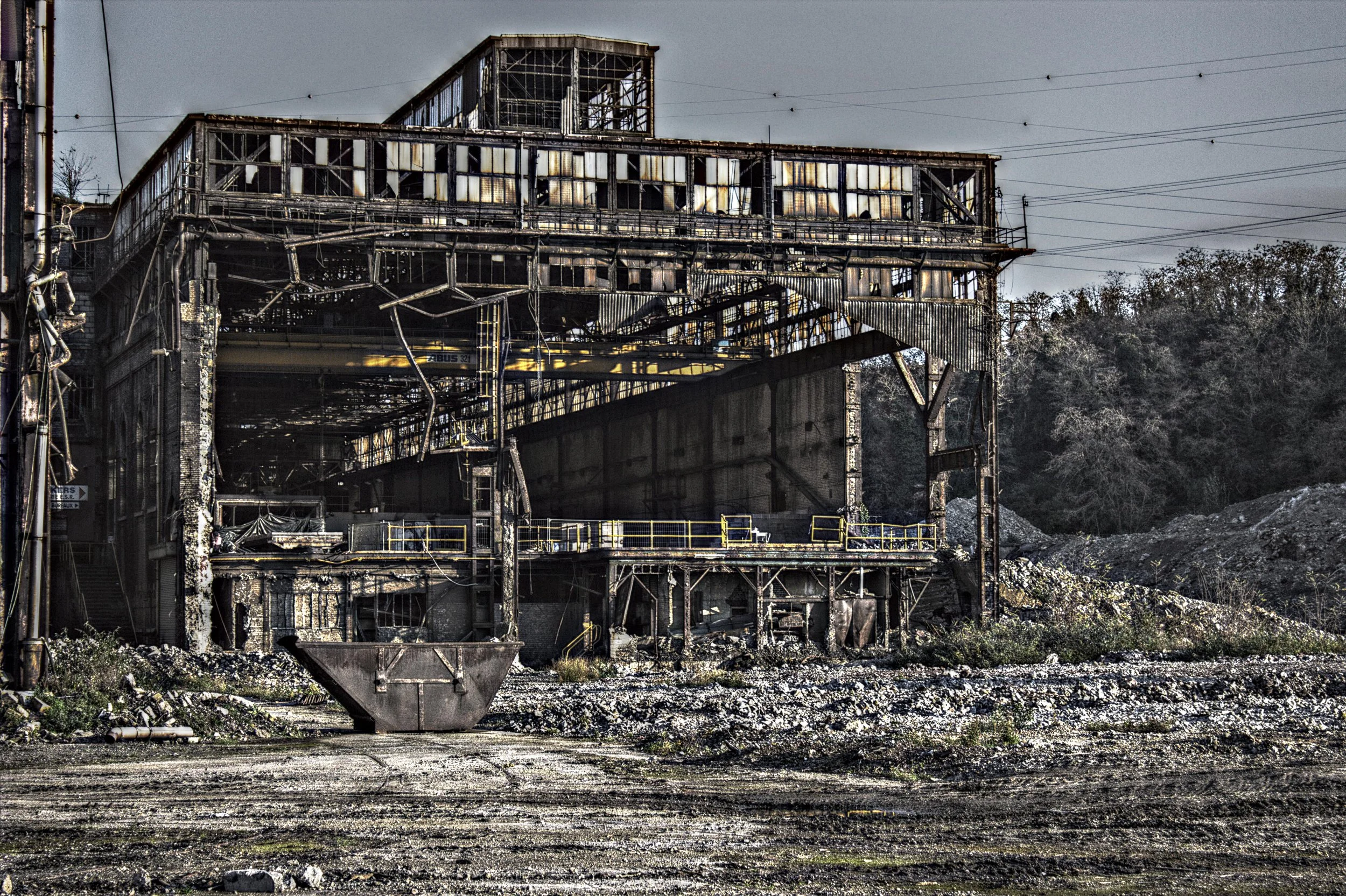 Verrostete, verlassene Industriehalle mit Zäunen, Kabeln und umgebendem Schutt, Landschaft im Hintergrund
