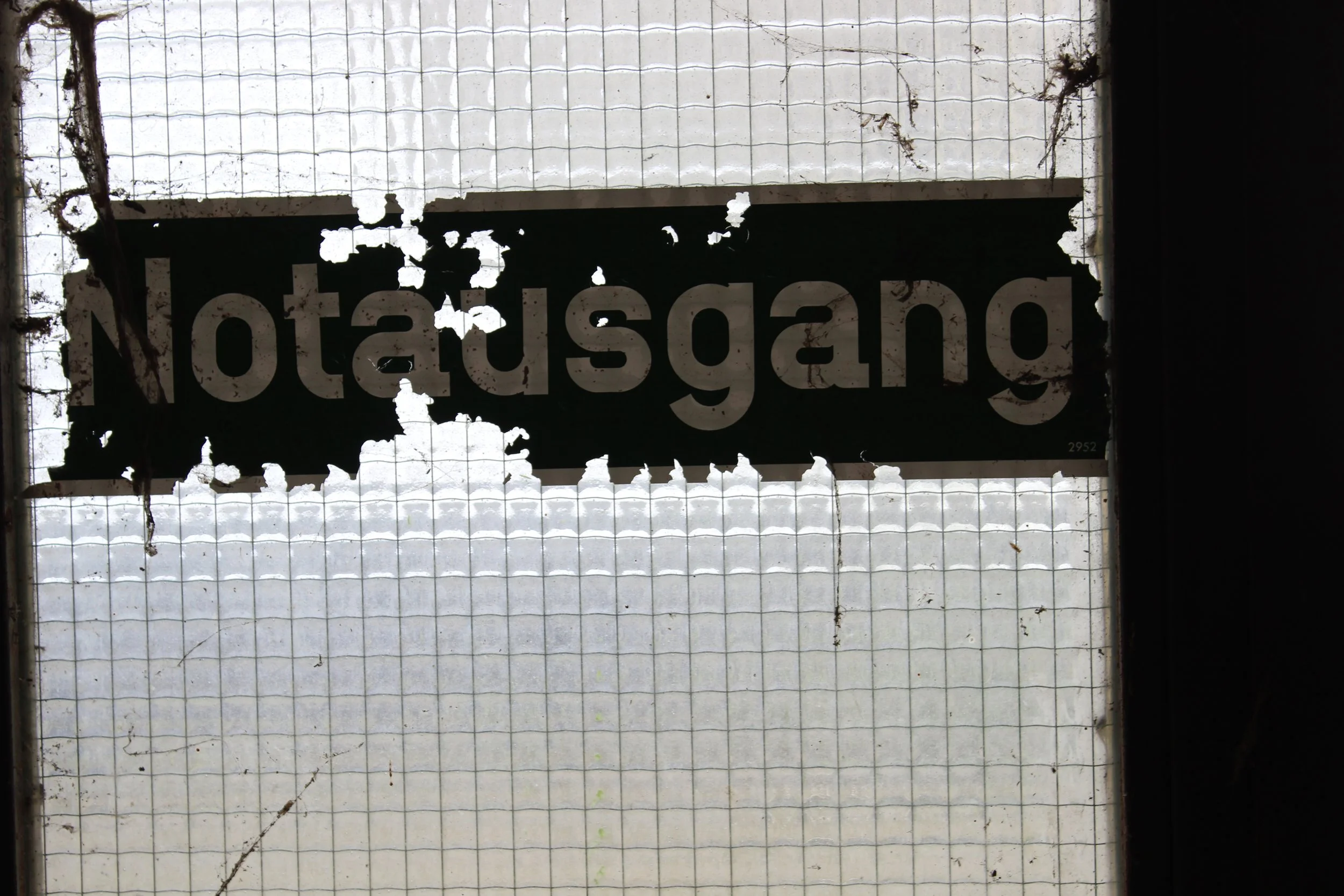 Verwittertes Schild mit dem deutschen Wort 'Notausgang', das an einer verschmutzten, zerbrochenen Scheibe hängt.