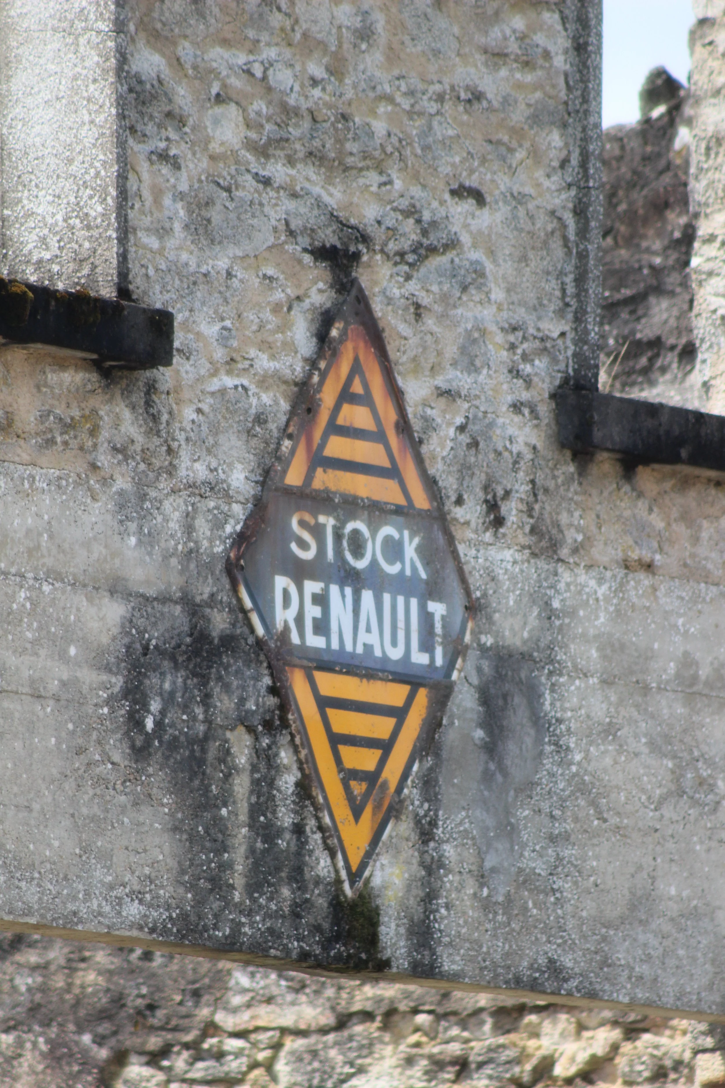 Einen alten, rostigen metallischen Schild an einer Steinmauer mit der Aufschrift "Stock Renault" und einem gelben Sicherheitszeichen.