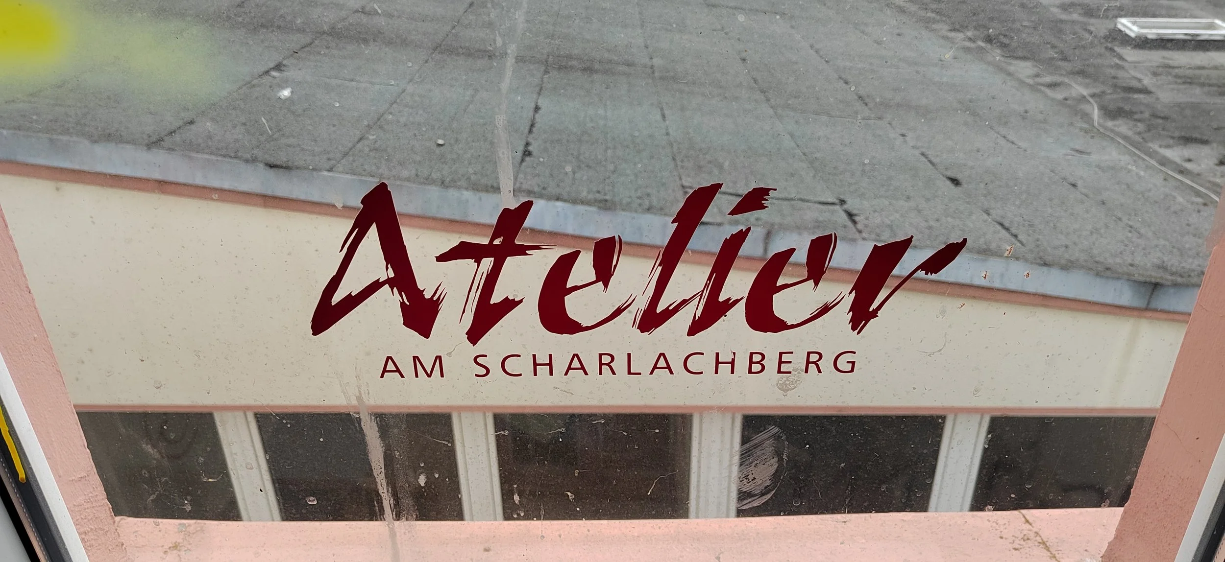 Hinweisschild mit der Aufschrift 'Atelier am Scharlachberg' auf einer Fensterscheibe, Blick auf die Straße und den Gehweg vor dem Atelier.