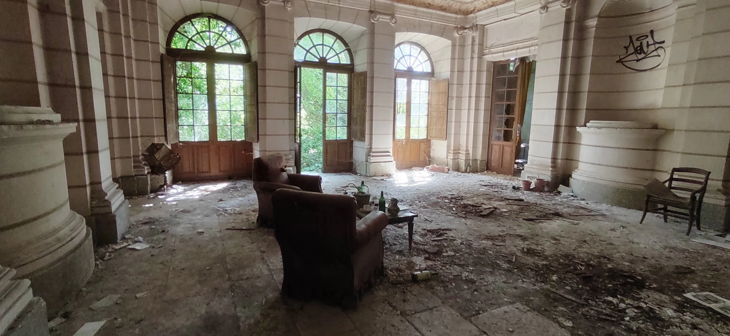 Verlassener, schmutziger Raum in einem alten Gebäude mit großen Fenstern, kaputten Türen, zerzaustem Boden und Möbeln wie Stühlen und einer Couch, die mit leeren Flaschen und Müll bedeckt sind.