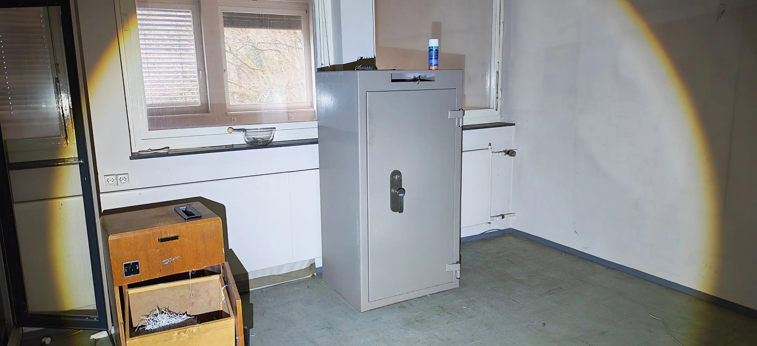 Leeres Zimmer mit Fenster, einer Gefriertruhe, einem alten Schrank, einer Glas-Schüssel auf dem Fensterladen und einem kleinen Holzschrank ohne Tür.