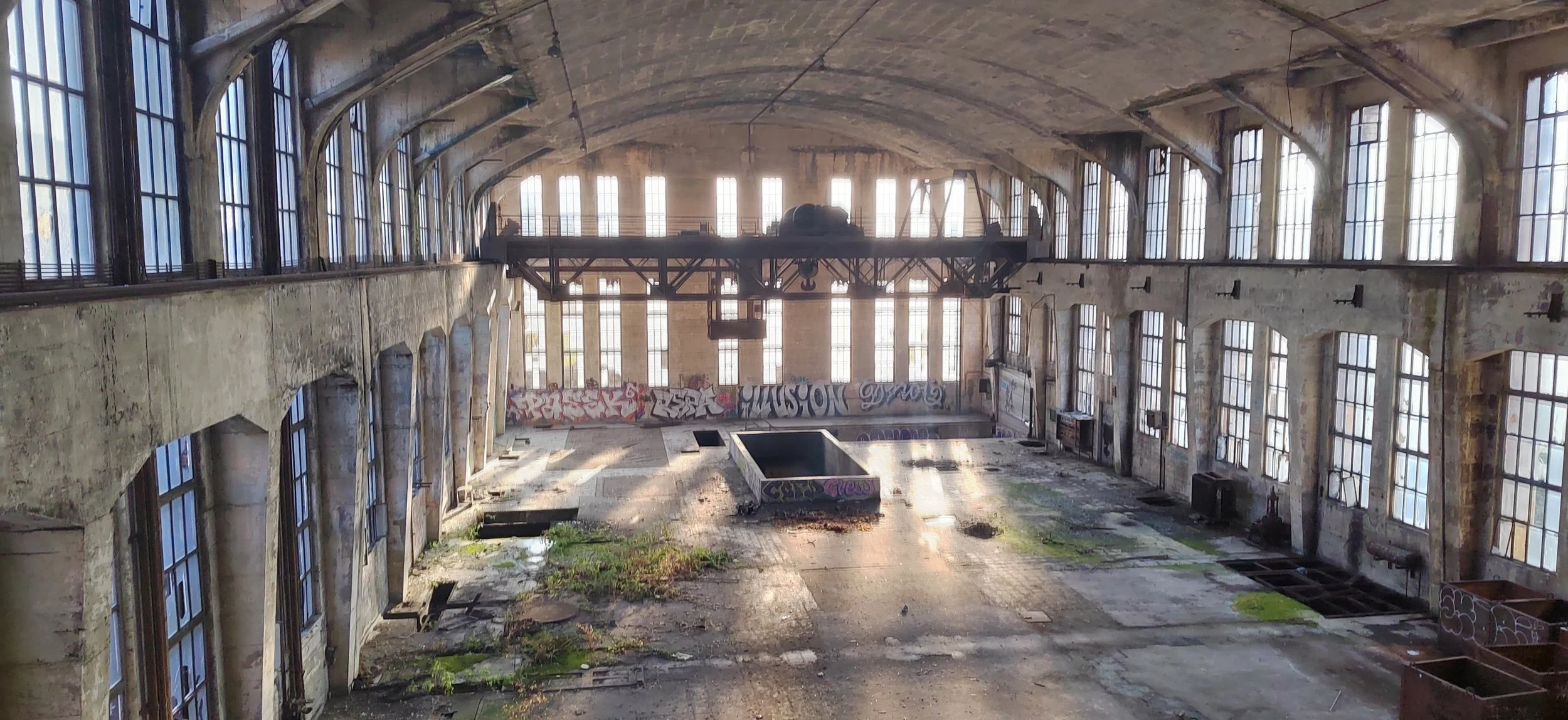 Ein verlassener, graffiti-bedeckter Turnhallenraum mit großen Fenstern und bröckelnder Wand- und Bodenstruktur.
