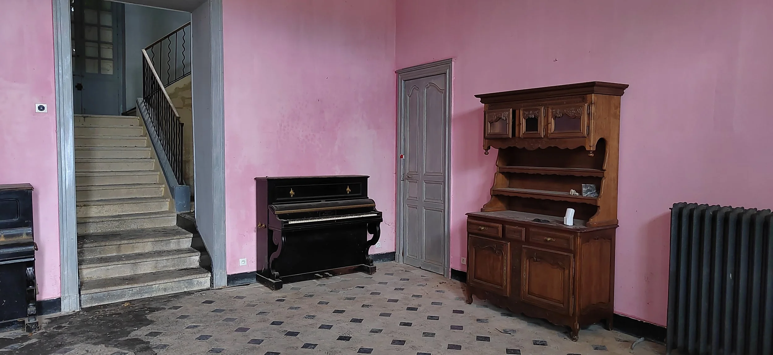 Ein Zimmer mit pinkfarbenen Wänden, einer alten schwarzen Klavier, einem Holzschrank und einer Heizungsanlage. Im Hintergrund eine Treppe mit steinernen Stufen und einem schwarzen Geländer.