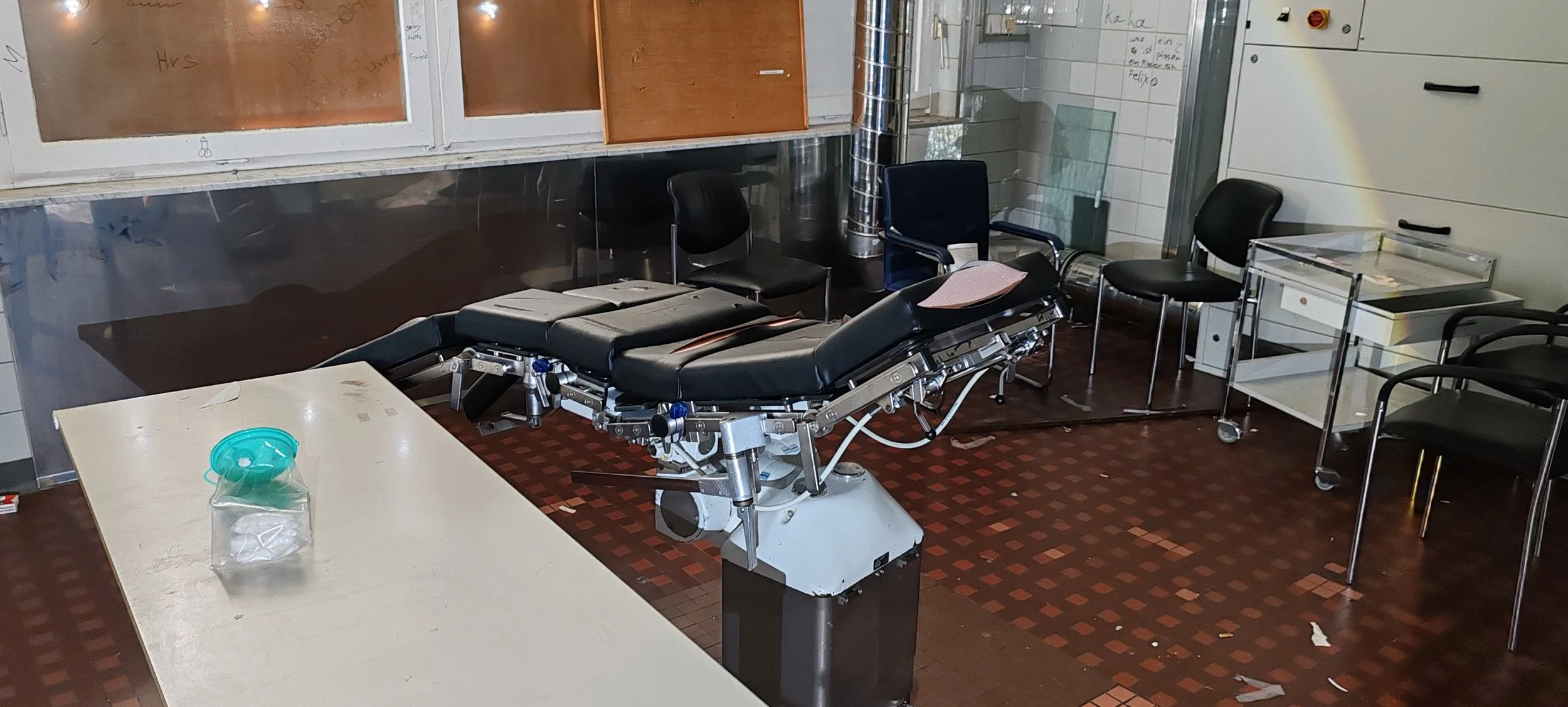 Ein leerer Operationsstuhl in einem Behandlungsraum, umgeben von schwarzen Stühlen und einem weißen Tisch mit einem Einweggesichtsschutz, im Hintergrund Fenster, Whiteboards und medizinische Geräte.