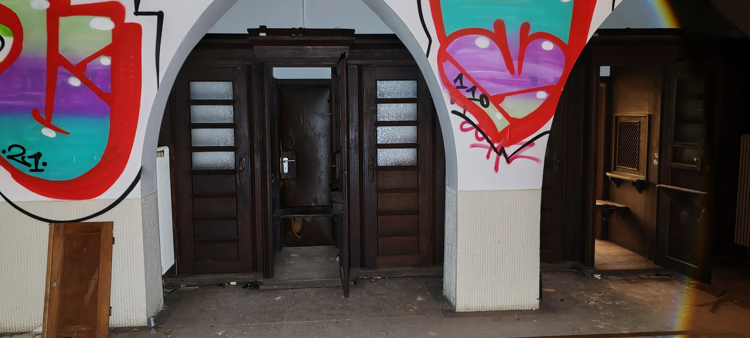 Innenraum eines verlassenen Gebäudes mit dunklen Holztüren und bunter Wandmalerei, Graffiti und Schmutz am Boden.