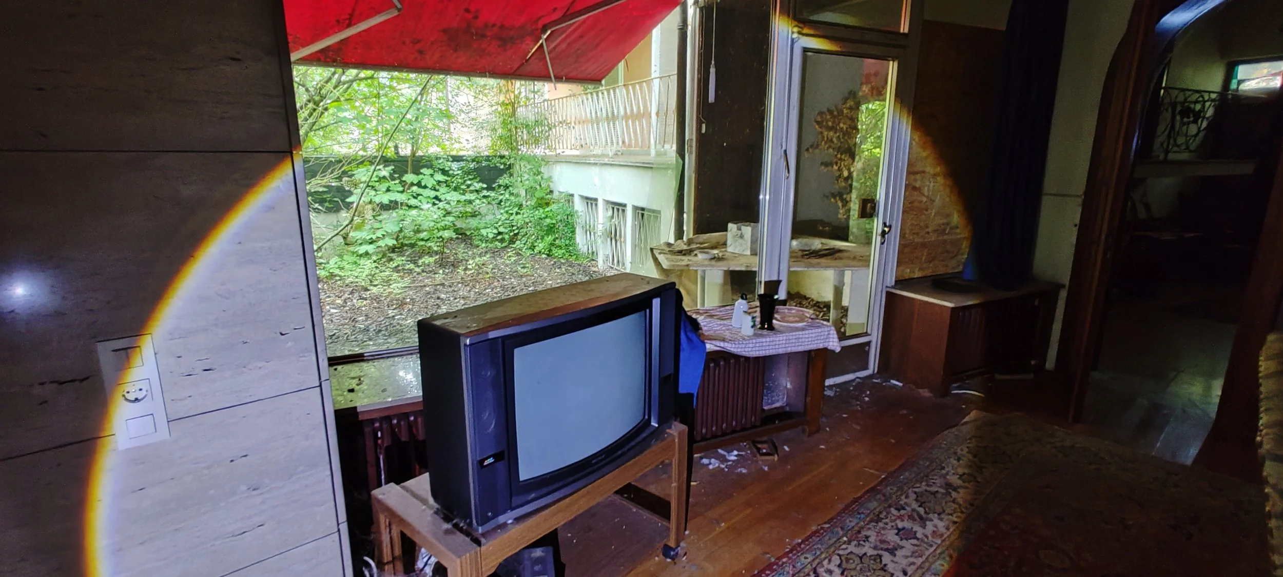 Ein Saal mit einem alten Fernseher auf einem Tisch, im Hintergrund ein offenes Fenster mit einer grünen Vogelvoliere und eine Tür, die auf einen bewachsenen Außenbereich führt. Der Raum wirkt ungepflegt und ungeordnet.
