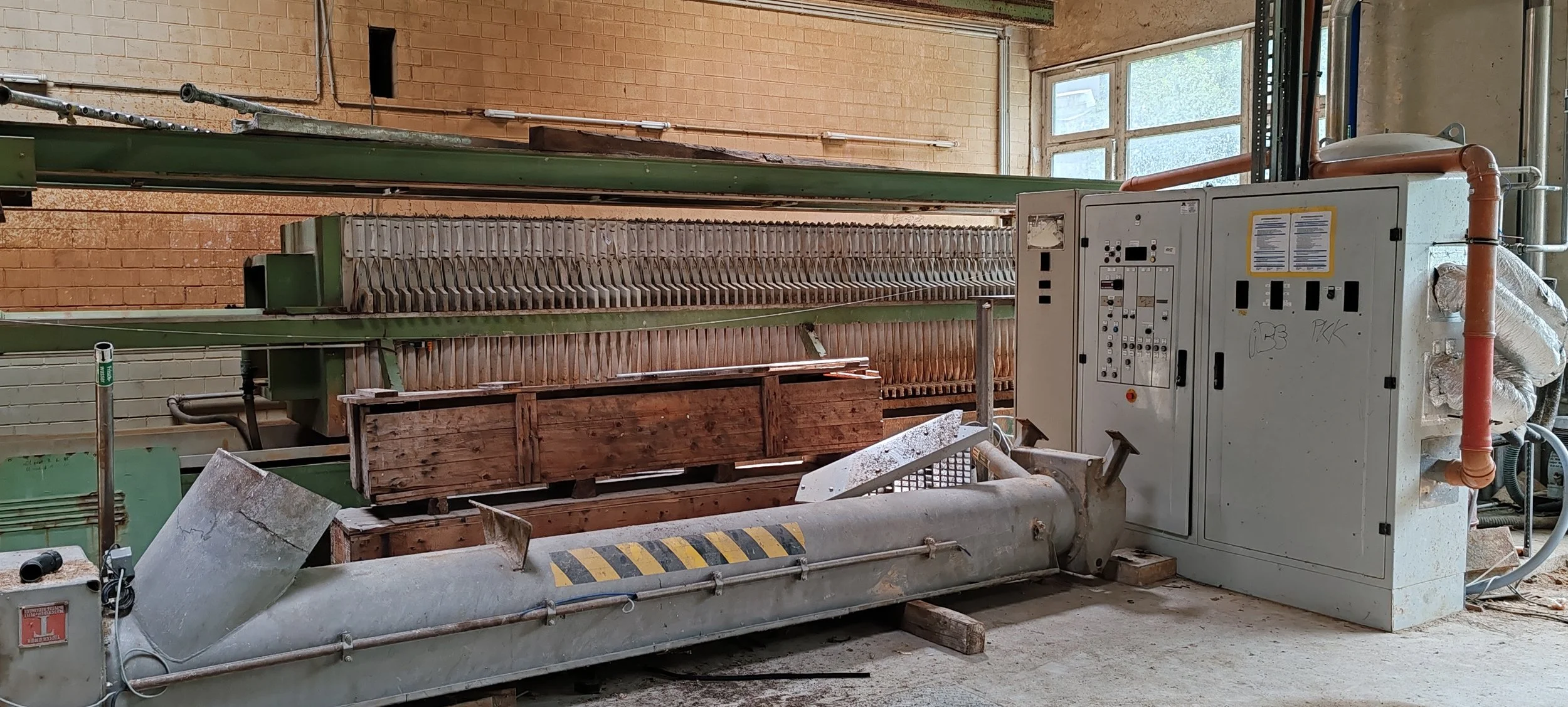 Industriemaschine in einer Werkstatt, mit Holzresten und Bedienfeld, in einem Raum mit großen Fenstern und Ziegelwänden.