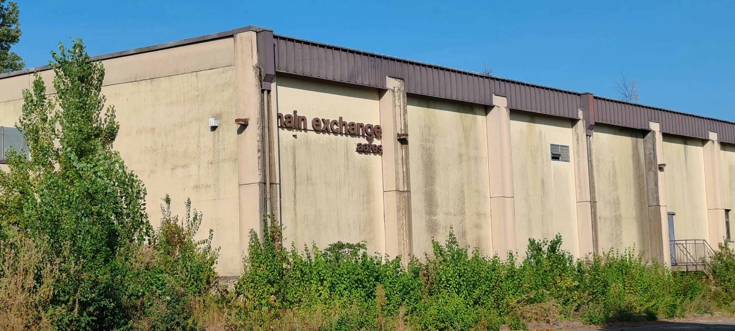 Außenansicht eines großen, grauen Gebäudes mit der Beschriftung "main exchange" an der Wand, umgeben von Büschen und Bäumen, bei sonnigem Himmel.