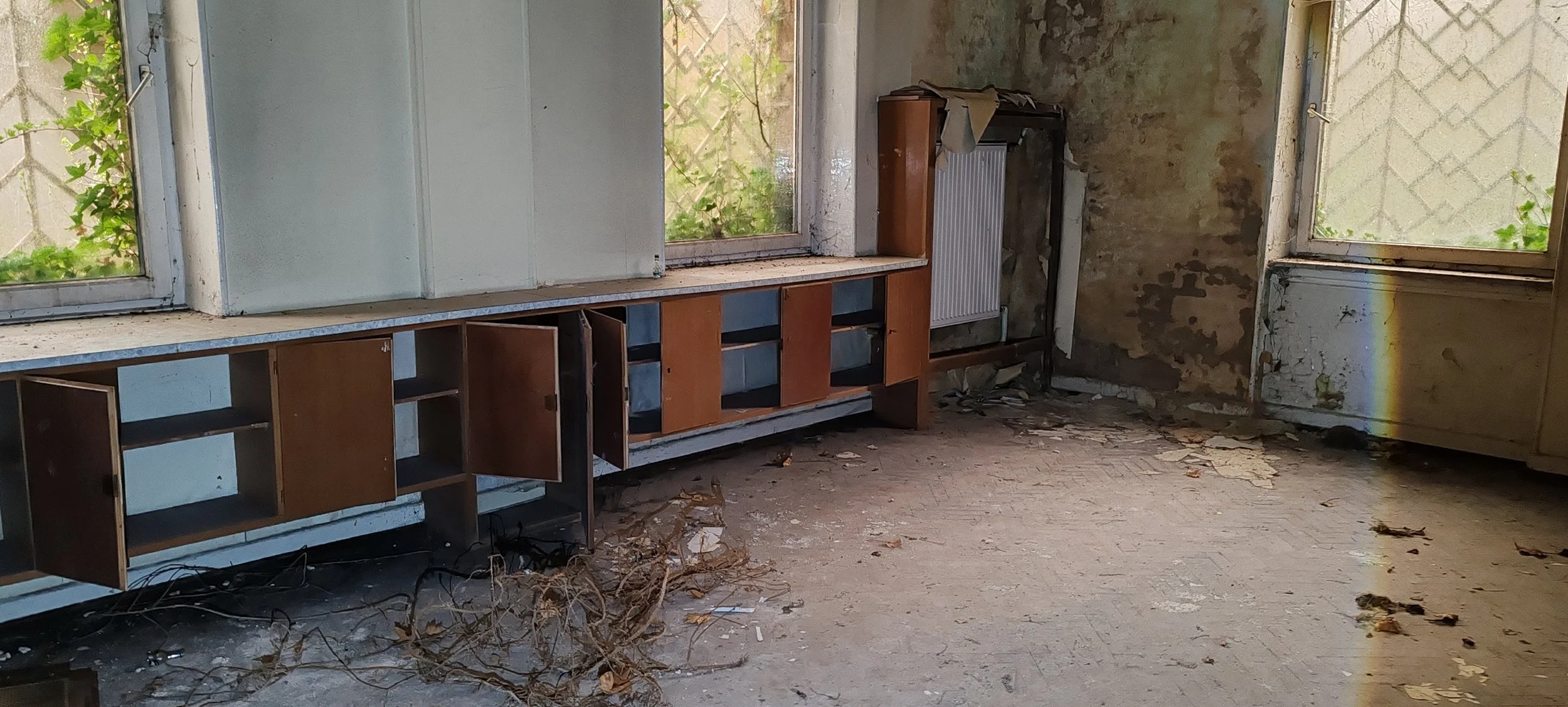 Verlassener, verfallener Raum mit kaputtem Boden, leeren Schränken und schmutzigen Wänden bei Tageslicht.