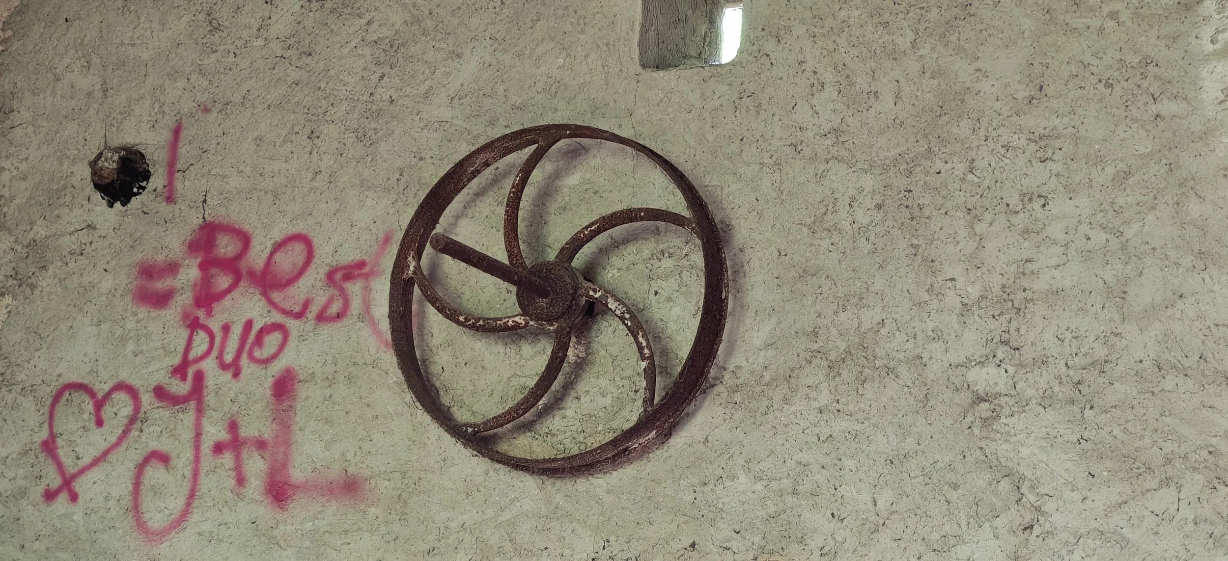 Ein alter, rostiger Ventilator auf einer hellen Wand mit pinker Graffiti-Schrift. Die Wand hat eine kleine, rechteckige Öffnung.