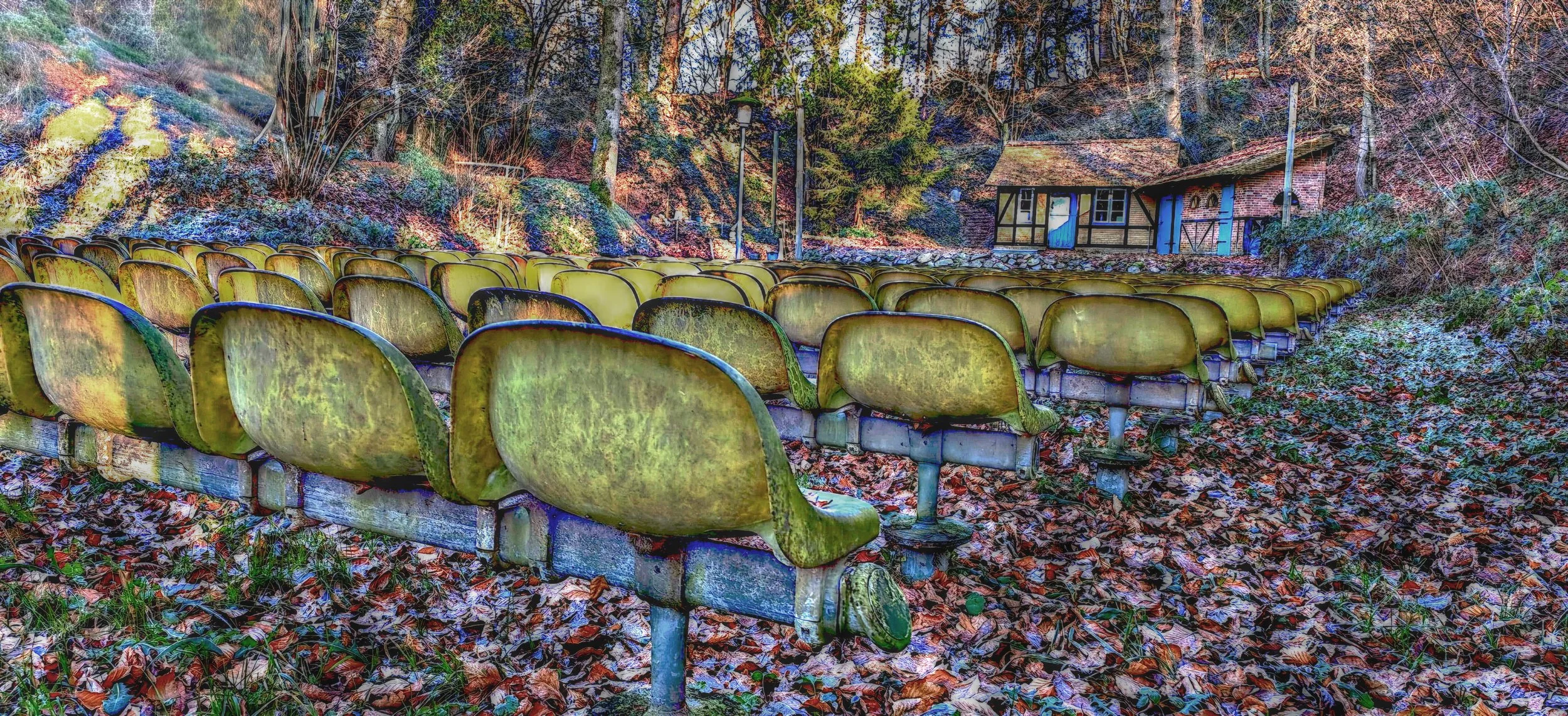 Verlassene, grüne, verwaschen aussehende Sitzbänke im Freien vor einem hügeligen, bewaldeten Hintergrund mit einem kleinen Fachwerkhaus im Hintergrund, umgeben von Herbstlaub.
