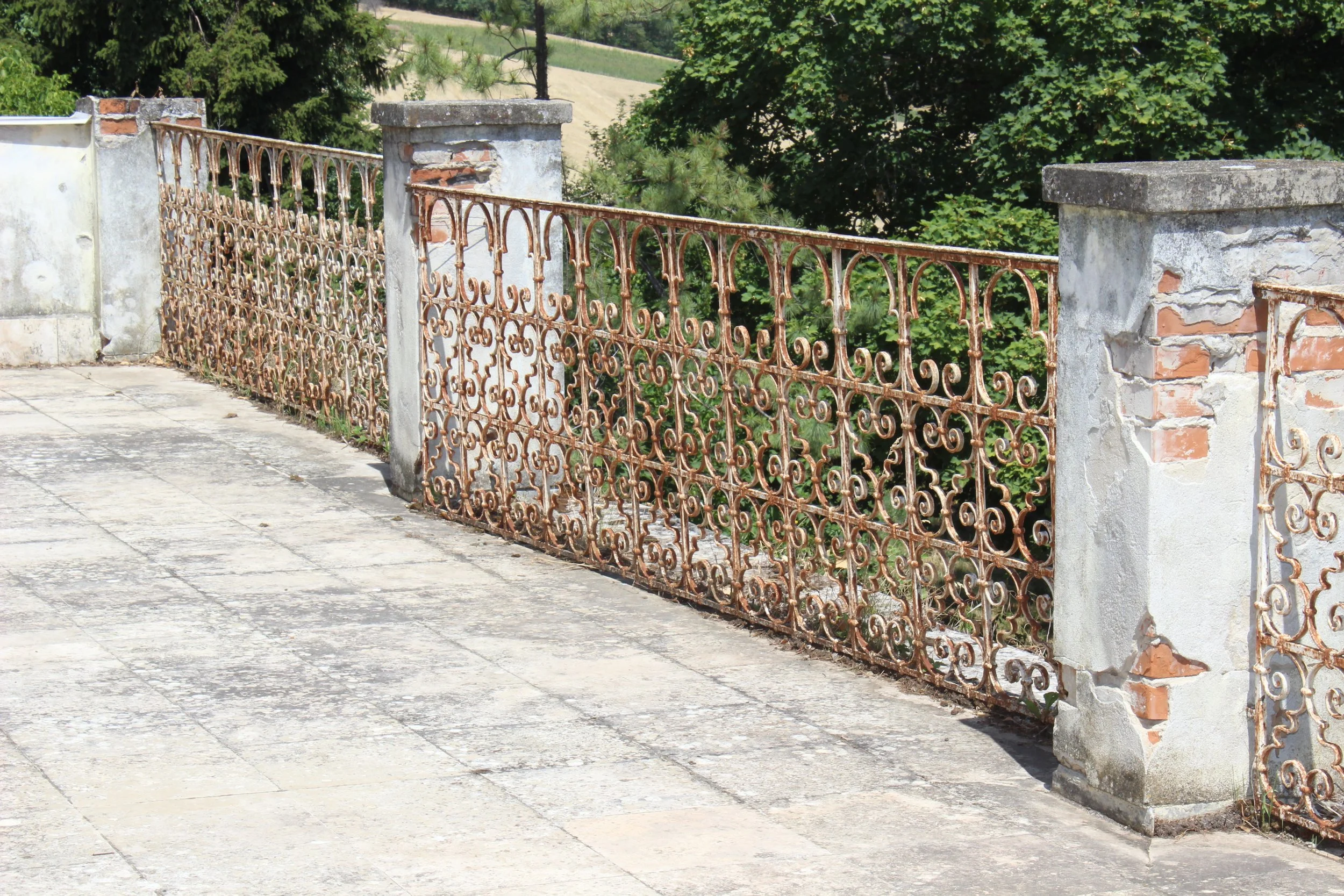 Eisenzaun an einem Balkon, mit Steinsäulen, darüber grüne Bäume.