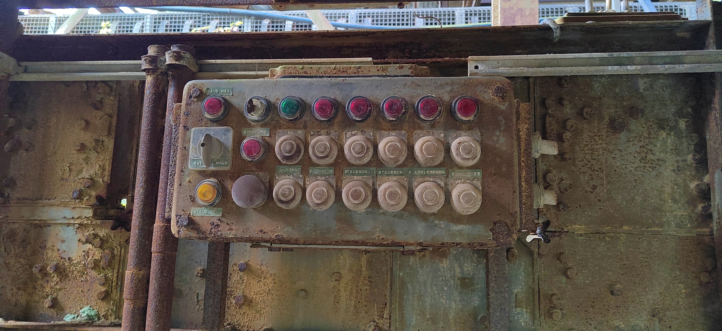 Alte, verrostete Steuerungs- oder Bedienbox aus Metall mit Kratzern, Staub und Rost, auf einem industriellen oder maschinellen Rahmen montiert.