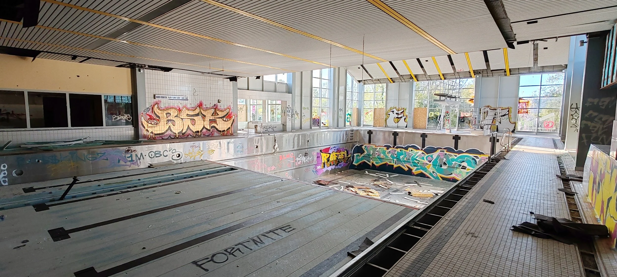 Leeres, verfallenes Schwimmbad mit Graffiti an den Wänden und Wasser im Becken, das mit Holz und Müll bedeckt ist.