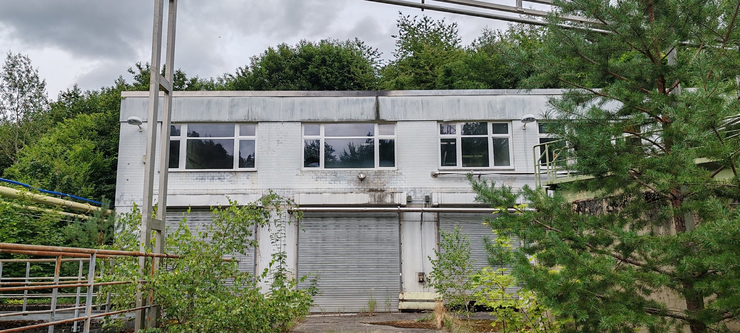 Ein verfallenes, zweistöckiges Gebäude mit weißen Wänden und großen Fenstern, umgeben von Bäumen und Pflanzen, mit einem grauen, bewölkten Himmel im Hintergrund.