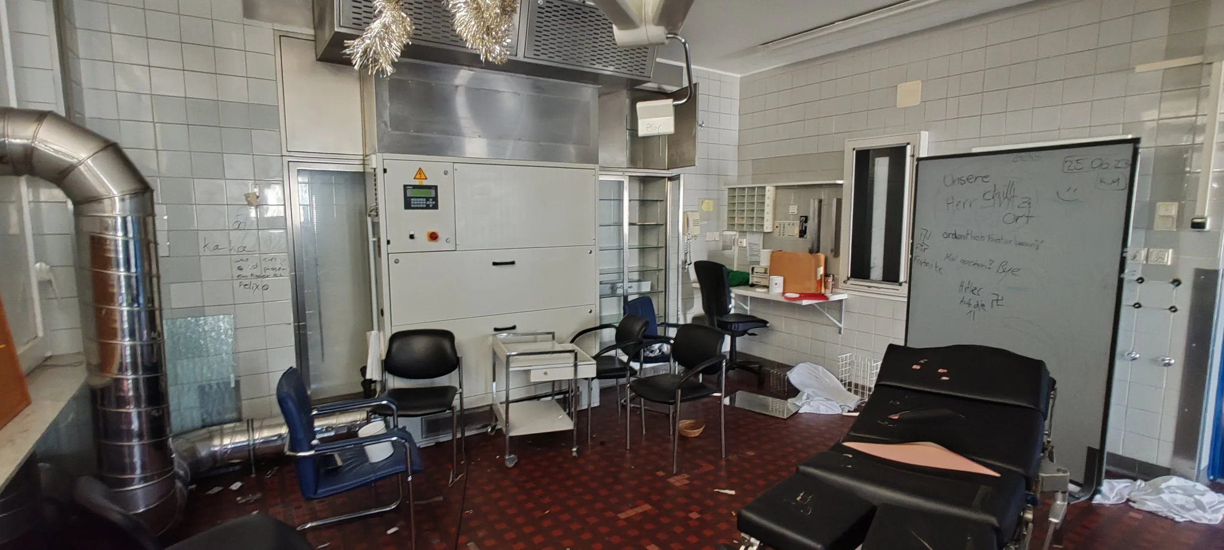 Ein chaotischer Raum mit mehreren schwarzen und blauen Stühlen, einem Whiteboard mit Schreibarbeiten, einem Fenster, einem Tisch mit Akten, einem weißen Schrank und einem schwarzen Behandlungsstuhl.
