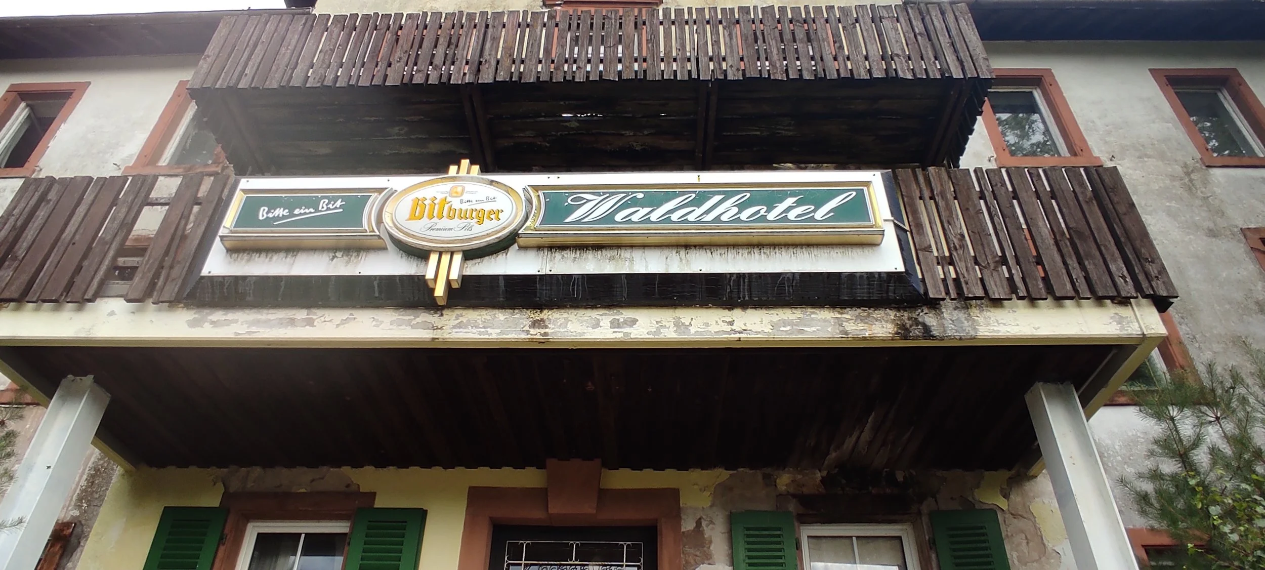Das Foto zeigt einen alten, an einer Fassade befestigten Balkon mit einer Holzbrüstung. Darunter befindet sich eine Ladenfront mit einem auffälligen Schild, das für ein Hotel namens "Valdhotel" wirbt, mit einem zusätzlichen Logo für "Bitburger" und der Aufforderung "Bitte ein Bit".