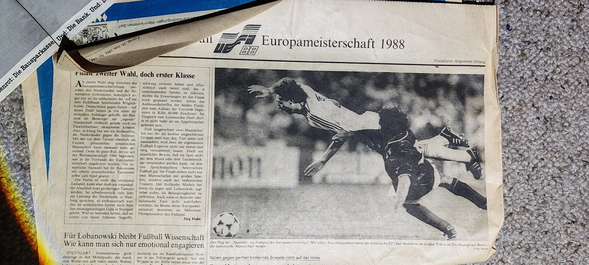 Schwarz-weiß-Fotografie eines Fußballspiels bei der Europameisterschaft 1988. Ein Spieler in einem weißen Trikot läuft, während ein anderer in einem dunklen Trikot fällt, mit einem Fußball auf dem Spielfeld.