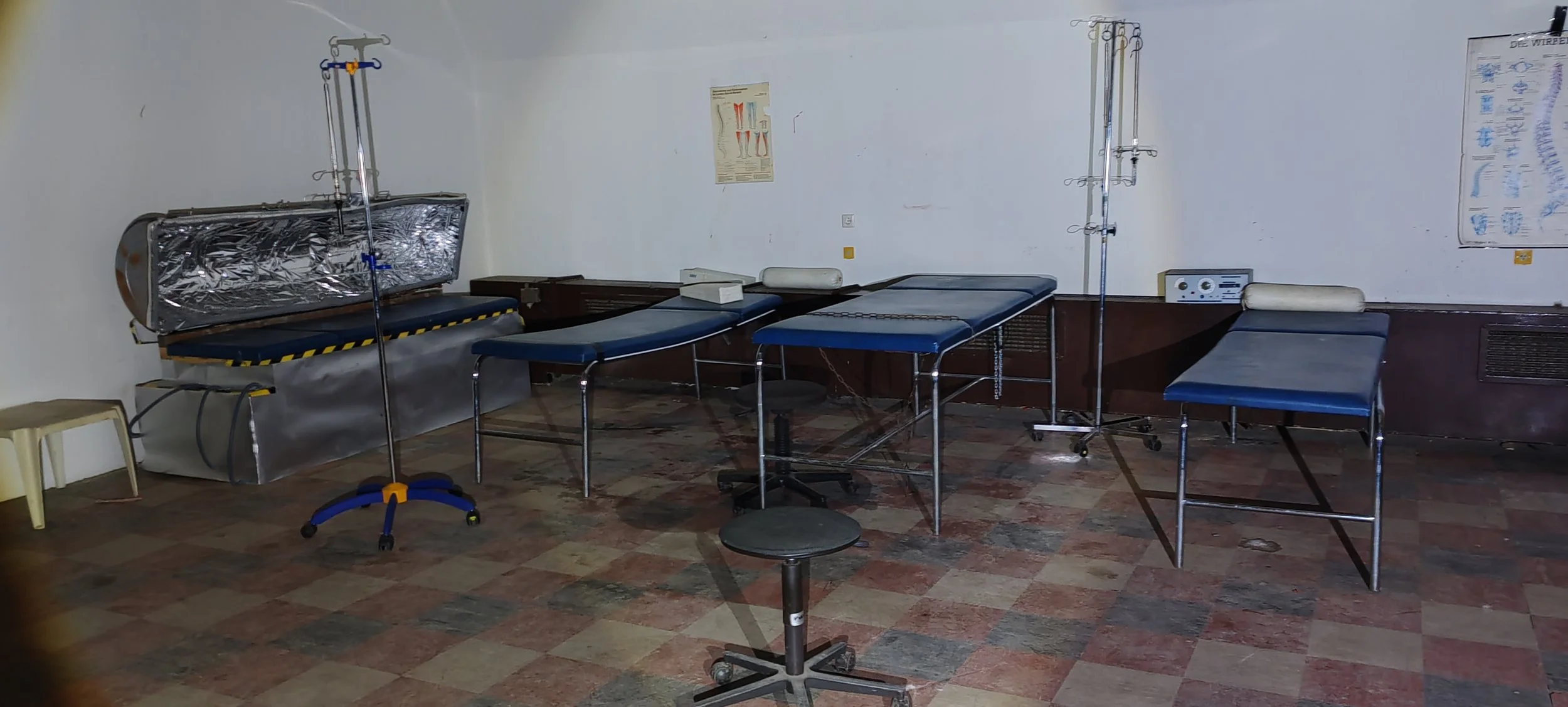Ein medizinischer Behandlungsraum mit mehreren Untersuchungsliegen, einem Rollenstuhl, Wänden mit medizinischen Diagrammen und medizinischen Geräten.