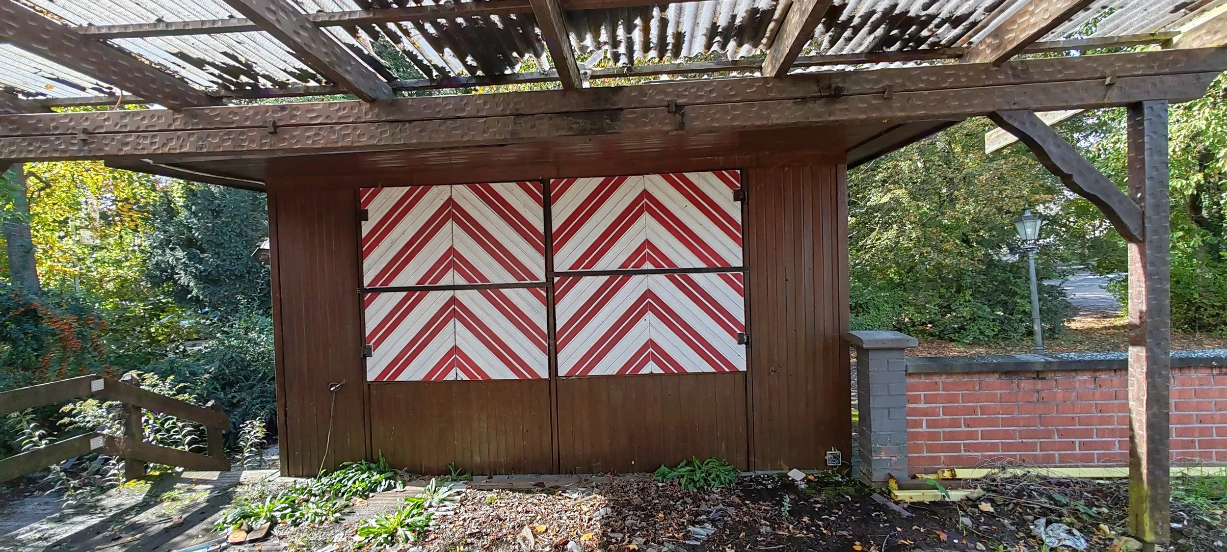 Holzschuppen mit rot-weißen bemalten doppelläufigen Türen, umgeben von Bäumen und einen Pflastermauer auf der rechten Seite.