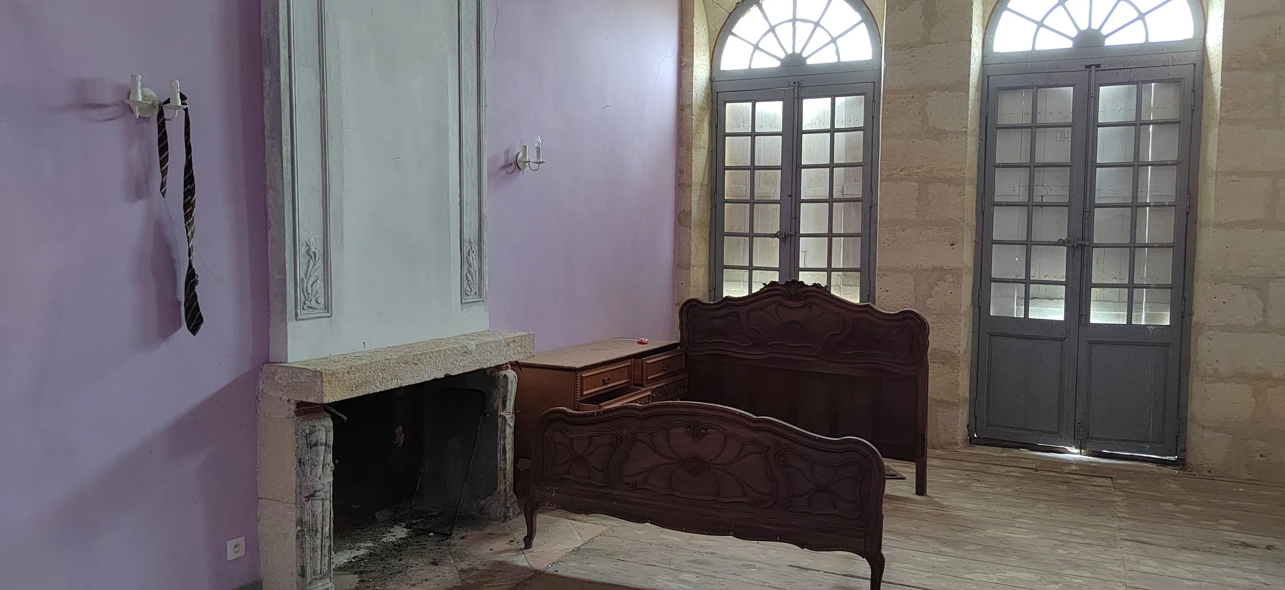 Ein Zimmer mit hohen Fenstern, Holzfußboden, einem alten Kamin und einem antiken Bett, die Wände sind hellviolett und beige.