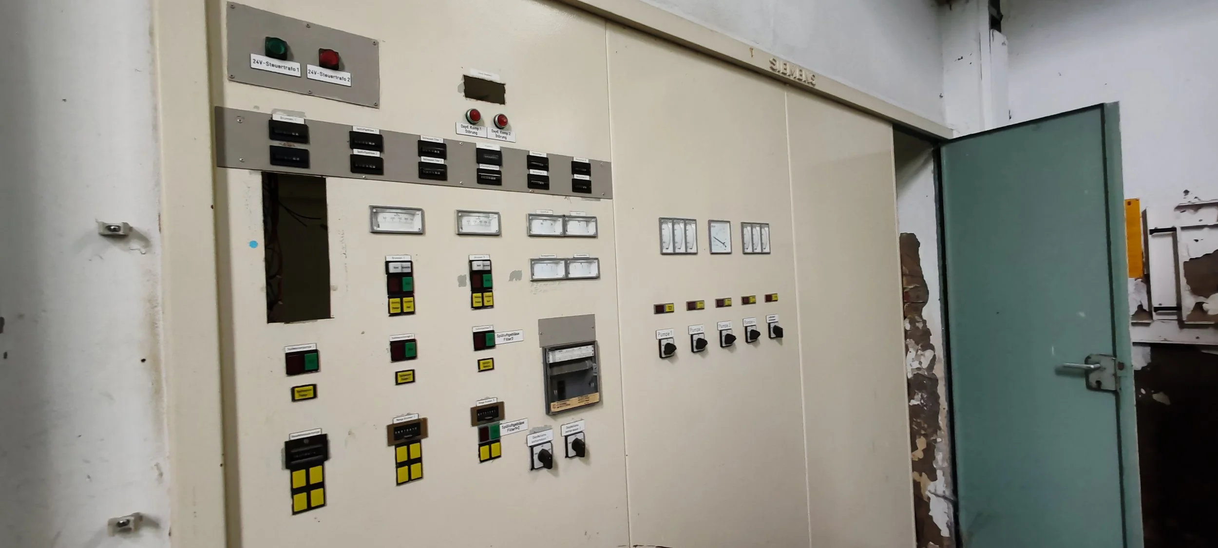 Alte elektrische Schalttafel mit verschiedenen Knöpfen, Anzeigen und Schaltern in einem Raum mit zerbrochener Wand und offenem grünen Tür.