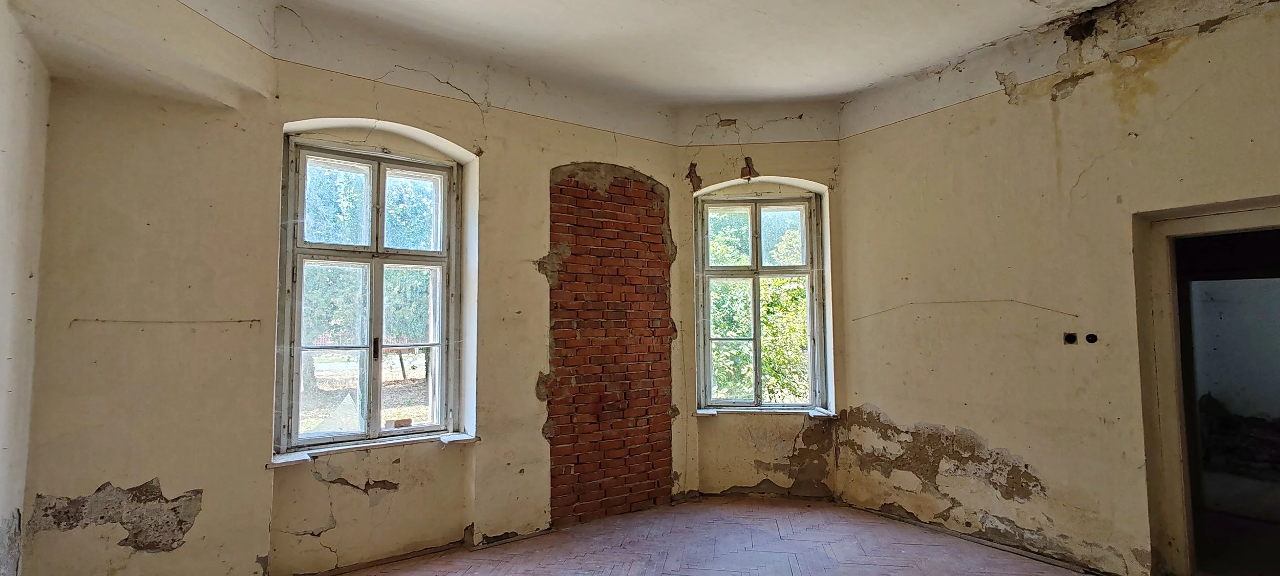 Ein alter, renovierungsbedürftiger Raum mit zwei Fenstern, an der Wand ist teilweise Putz abgefallen, und eine Wand wurde teilweise aus Ziegelsteinen freigelegt.