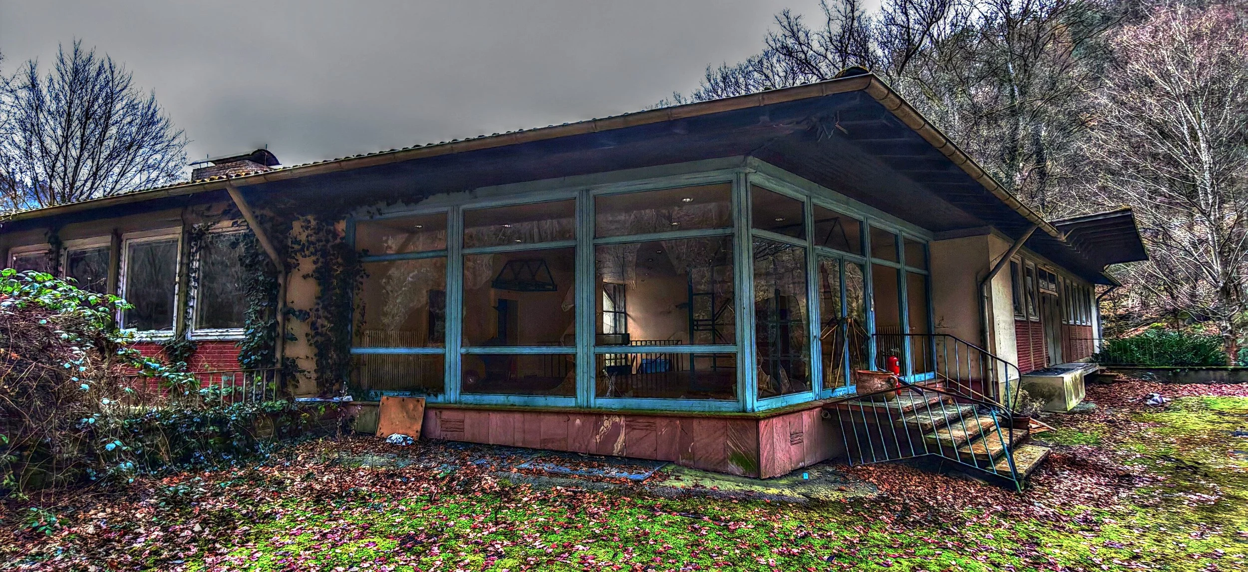 Verfallene, verlassene Haus mit großen Fenstern und bemaltem Unterbau, umgeben von Laub und Bäumen im Herbst.