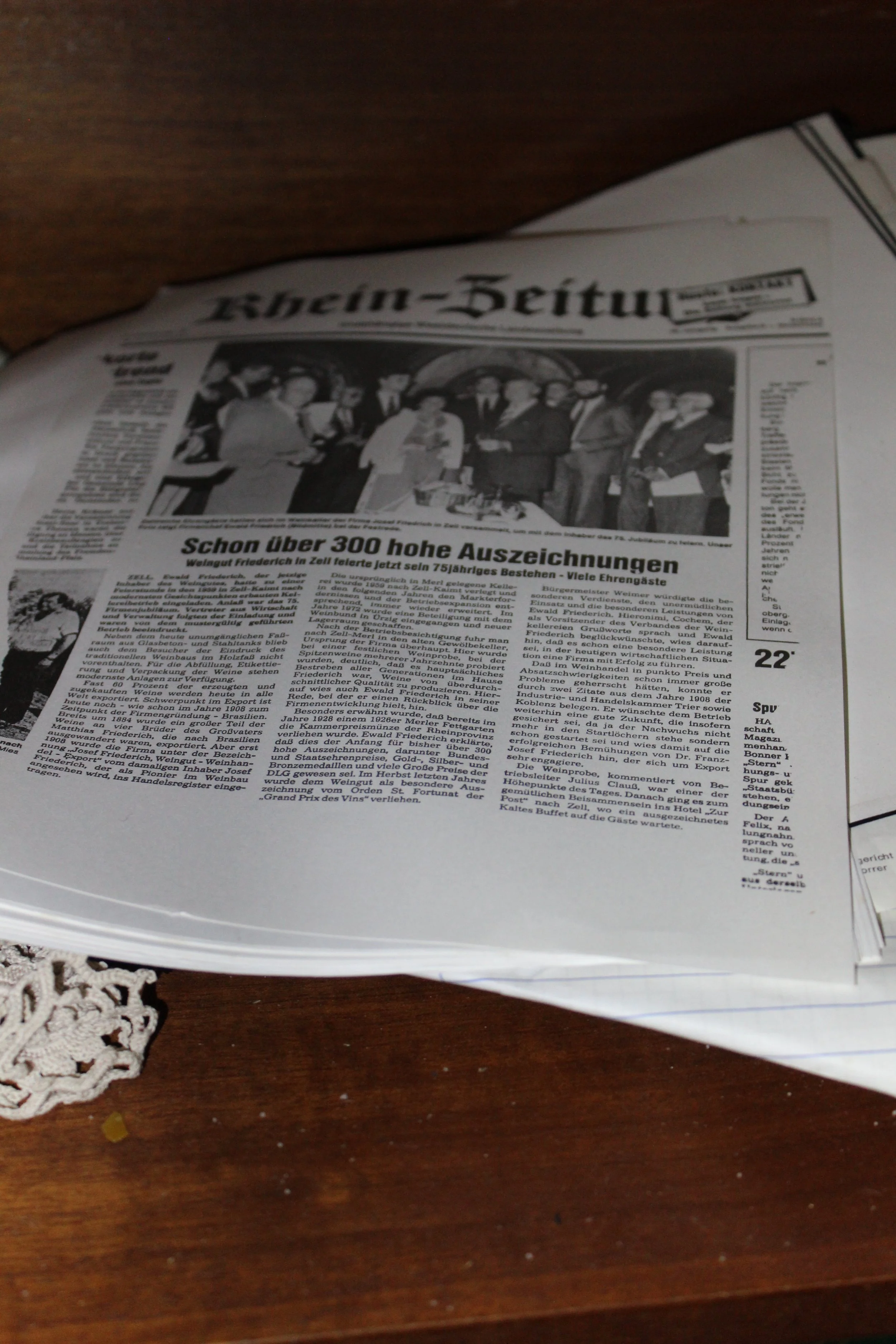 Zeitung 'Kreisin-Zeitung' liegt auf einem Holz Tisch, auf dem eine weiße Spitze liegt. Der Artikel berichtet über Auszeichnungen und Erfolge eines Weinbetriebs.