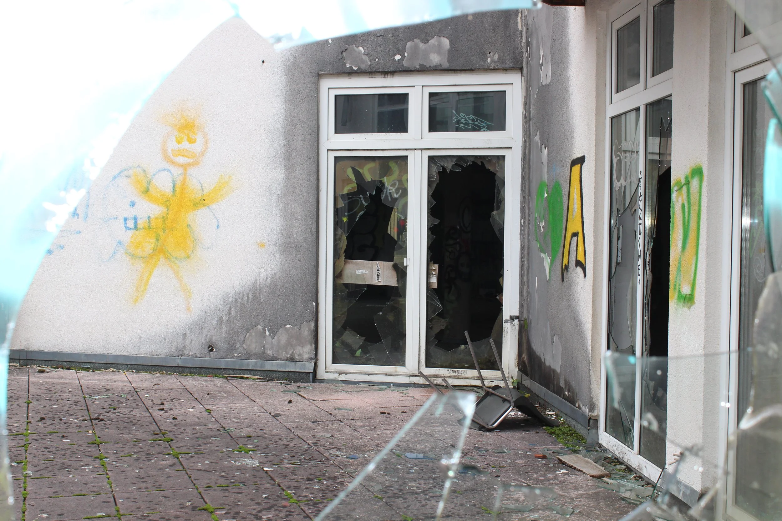 Ein zerstörtes Gebäude mit zerbrochenen Glasfenstern, Graffiti an den Wänden und umgestürztem Stuhl vor der Tür.