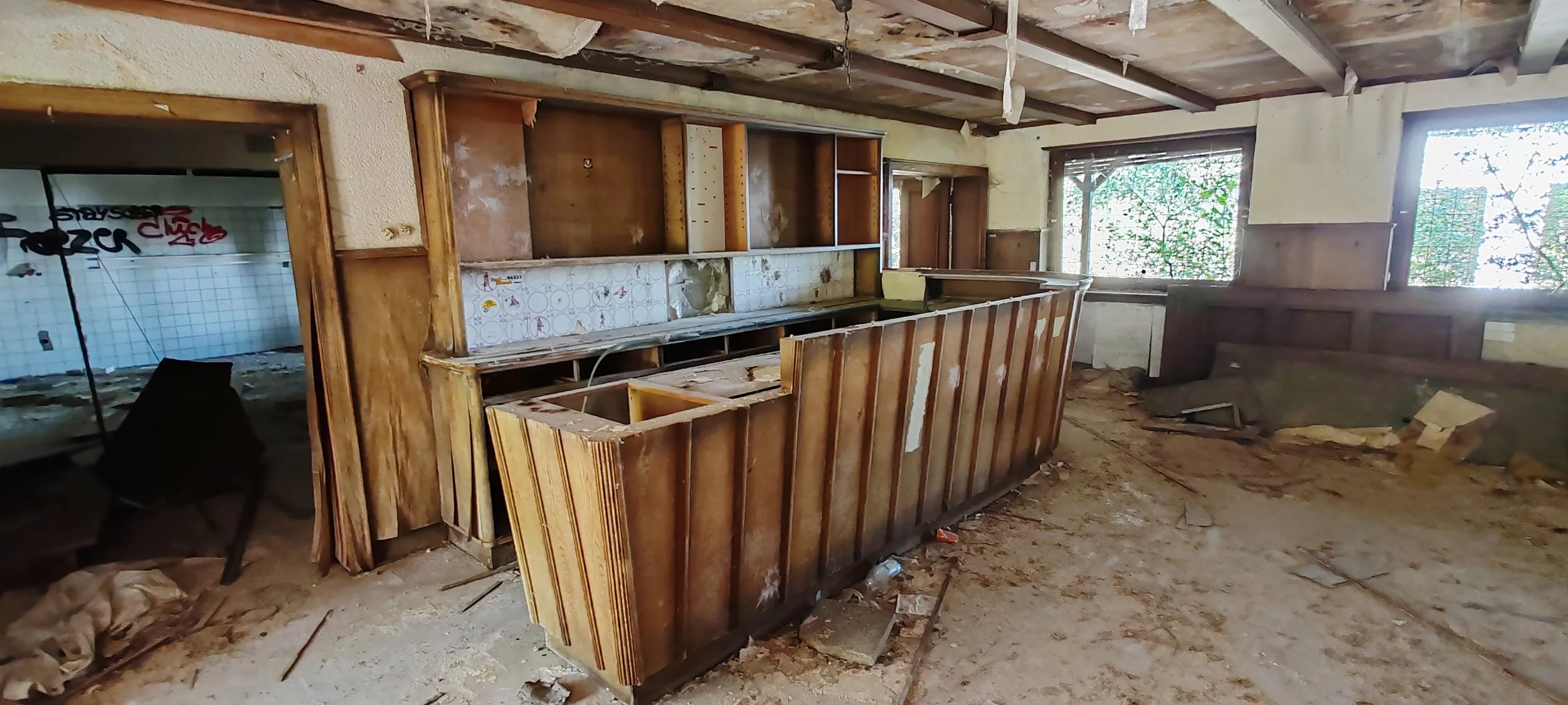 Verlassene, abgenutzte Bar mit beschädigter Holztheke und offenen Regalen in einem verfallenen Raum mit staubigem Boden und zerbrochenen Fenstern.