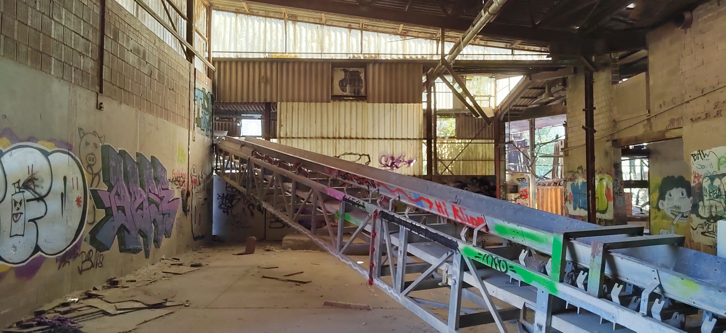 Verlassene, graffiti-beschmierte Industriehalle mit heruntergekommenem Gerüst und einigen kaputten Teilen, Sonnenlicht fällt durch Fenster. Kabel und Schutt auf dem Boden.