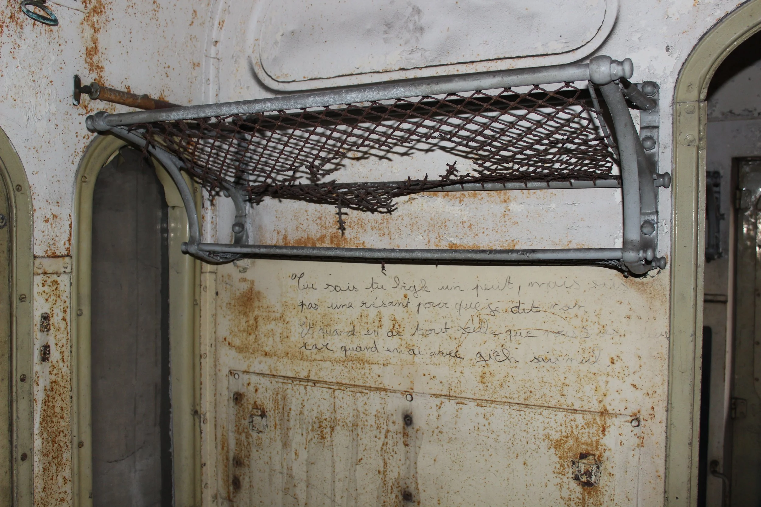 Verrosteter Metallschrank mit einem herausgenommenen, rustikalen Netztablett in einem alten, rostigen Raum, darunter handgeschriebene französische Notizen an der Wand.
