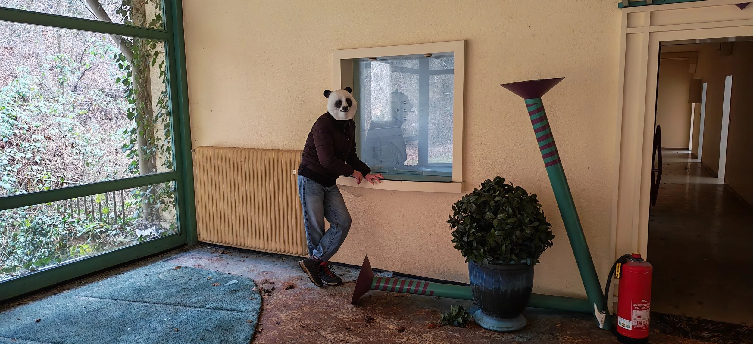 Person mit Panda-Maske steht in einem Flur, neben einem Fenster, auf einem grauen Teppich. Es gibt einen großen umgestürzten Blumentopf, eine grüne Pflanze, ein rotes Feuerlöscher und eine große Strickleiter. Im Hintergrund eine dunkle Tür und ein du