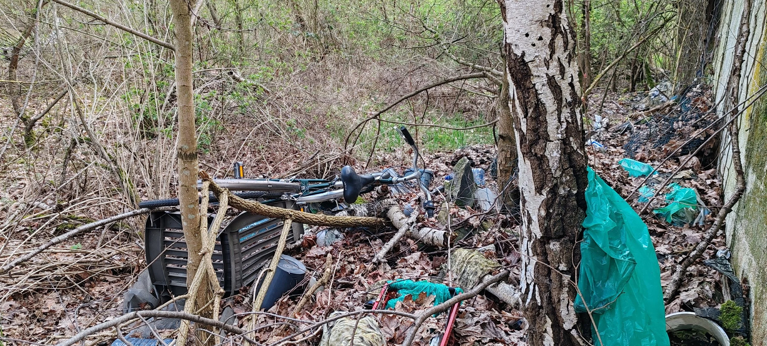 Verschmutzter Waldweg mit weggeworfenem Müll, darunter eine umgestürzte Fahrrad und Plastiktüten.