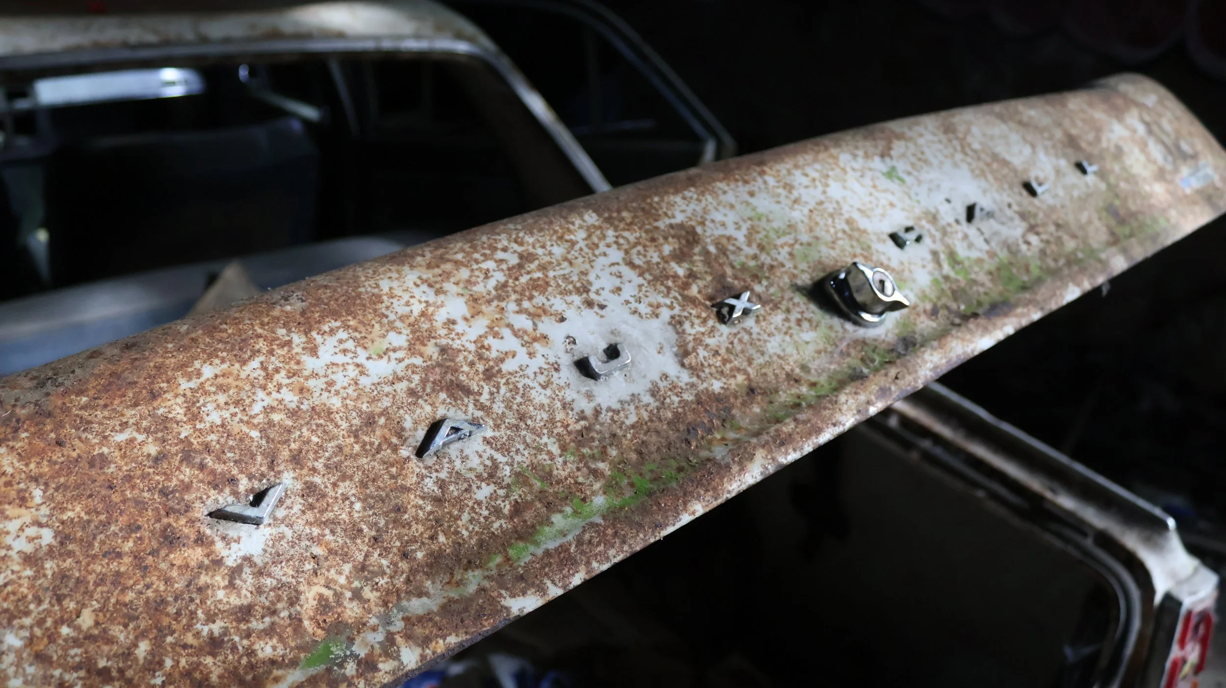 Ein verrostetes Metallteil mit alten Buchstaben, vermutlich von einem Autoblech, liegt auf einer Oberfläche. Im Hintergrund sind die Überreste eines Autos sichtbar.