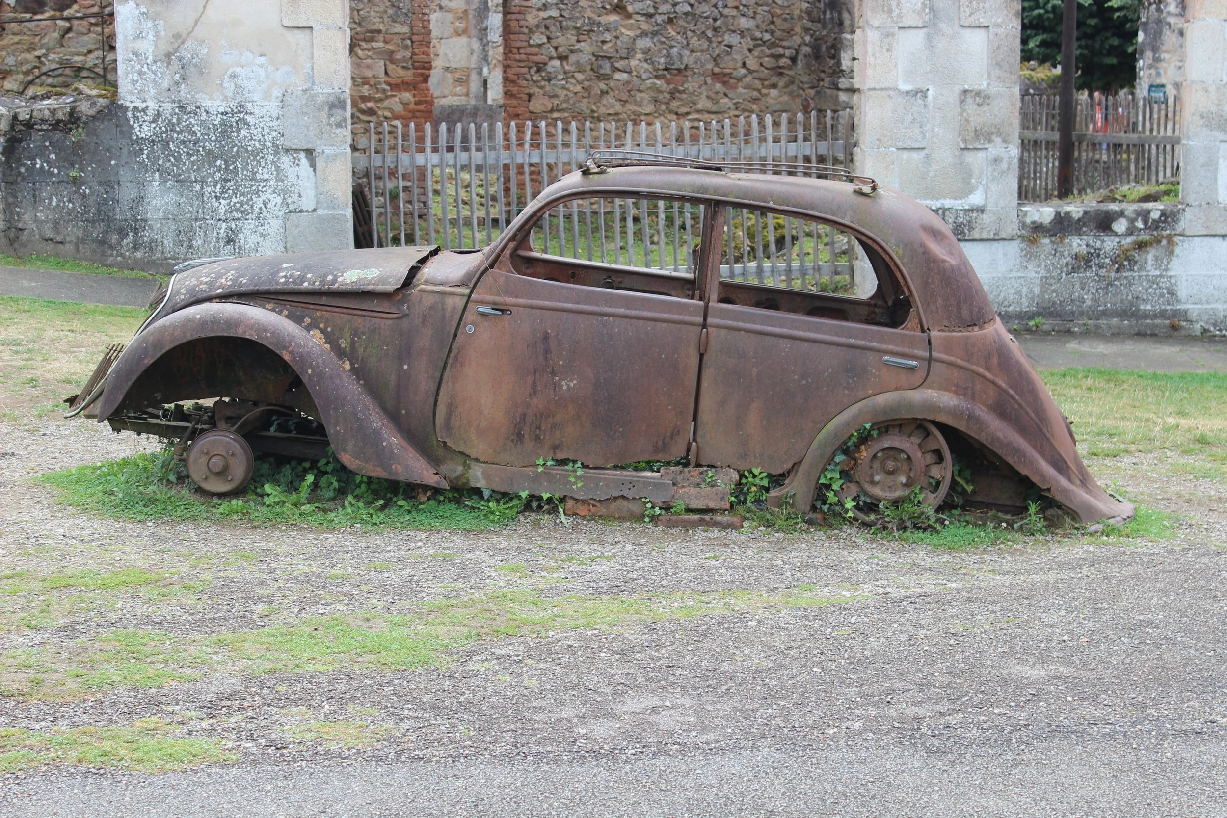 Ein verrosteter, alter Auto-Bauteil, das auf einem Boden liegt, ohne Räder, im Freien vor einer alten Steinmauer.