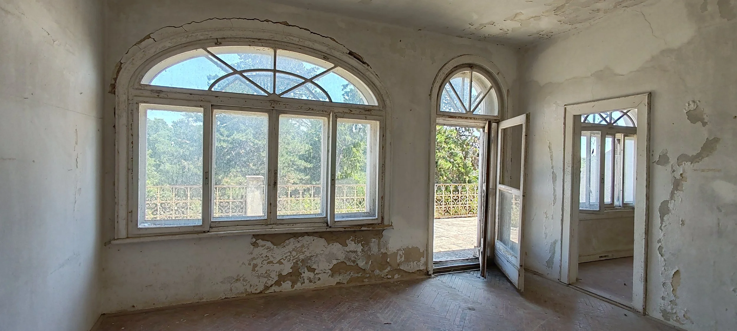 Ein verlassener, alter Raum mit großen Fenstern und Türen, Wände mit abblätternder Farbe, Blick nach draußen auf einen Balkon mit Geländer und Bäumen
