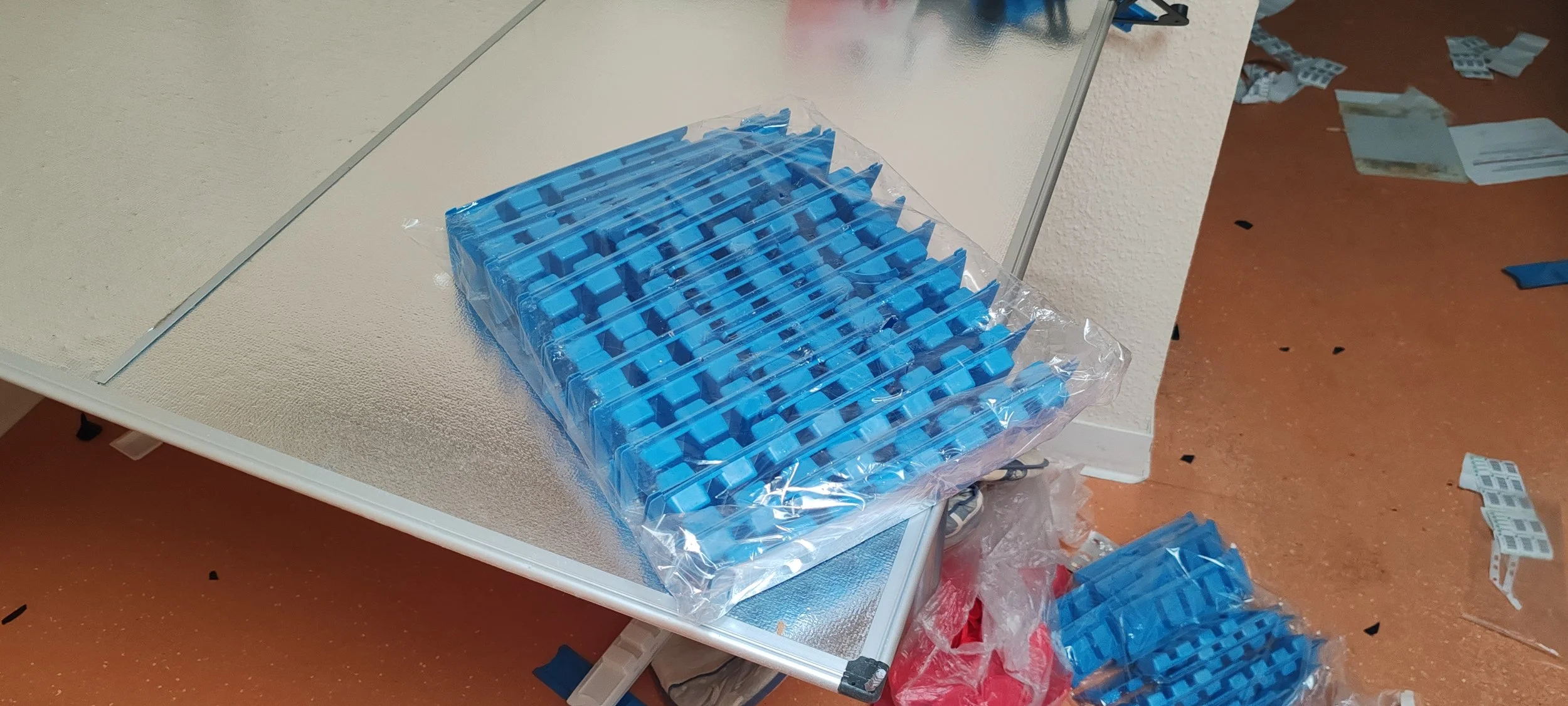 Blaue Kunststoffteile in einer Verpackung auf einem Tisch, umgeben von verstreuten Teilen auf dem Boden.