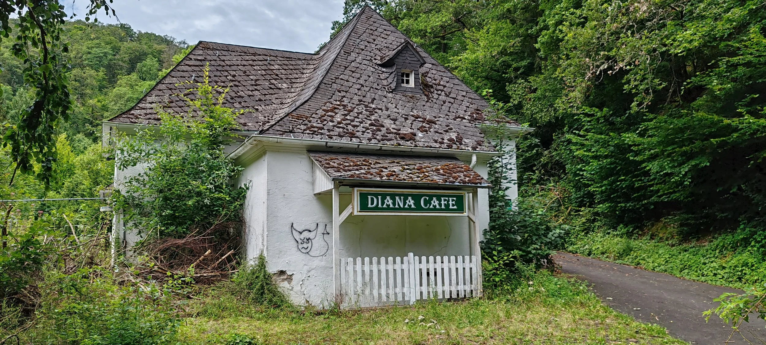 Ein kleines weißes Gebäude mit einem Schild, auf dem steht 'DIANA CAFE', liegt in einer grünen, bewaldeten Umgebung. Das Dach ist schief und mit Moos bedeckt. Es gibt ein kleines, schräges Fenster im Dach; an der Seite des Gebäudes ist eine einfache 