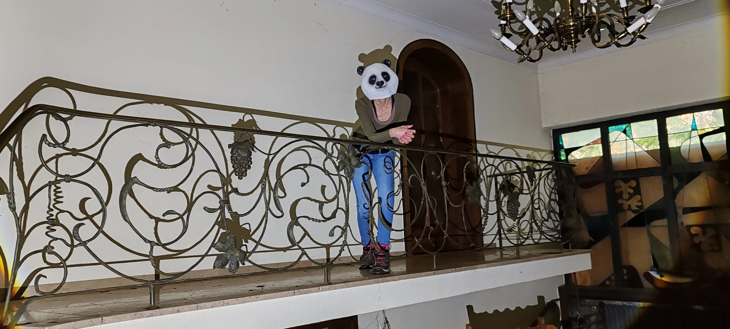 Person in Panda-Maske steht auf der Galerie eines Hauses mit verspieltem Eisenreling, im Hintergrund ein Kronleuchter und Fenster mit buntem Glas. 