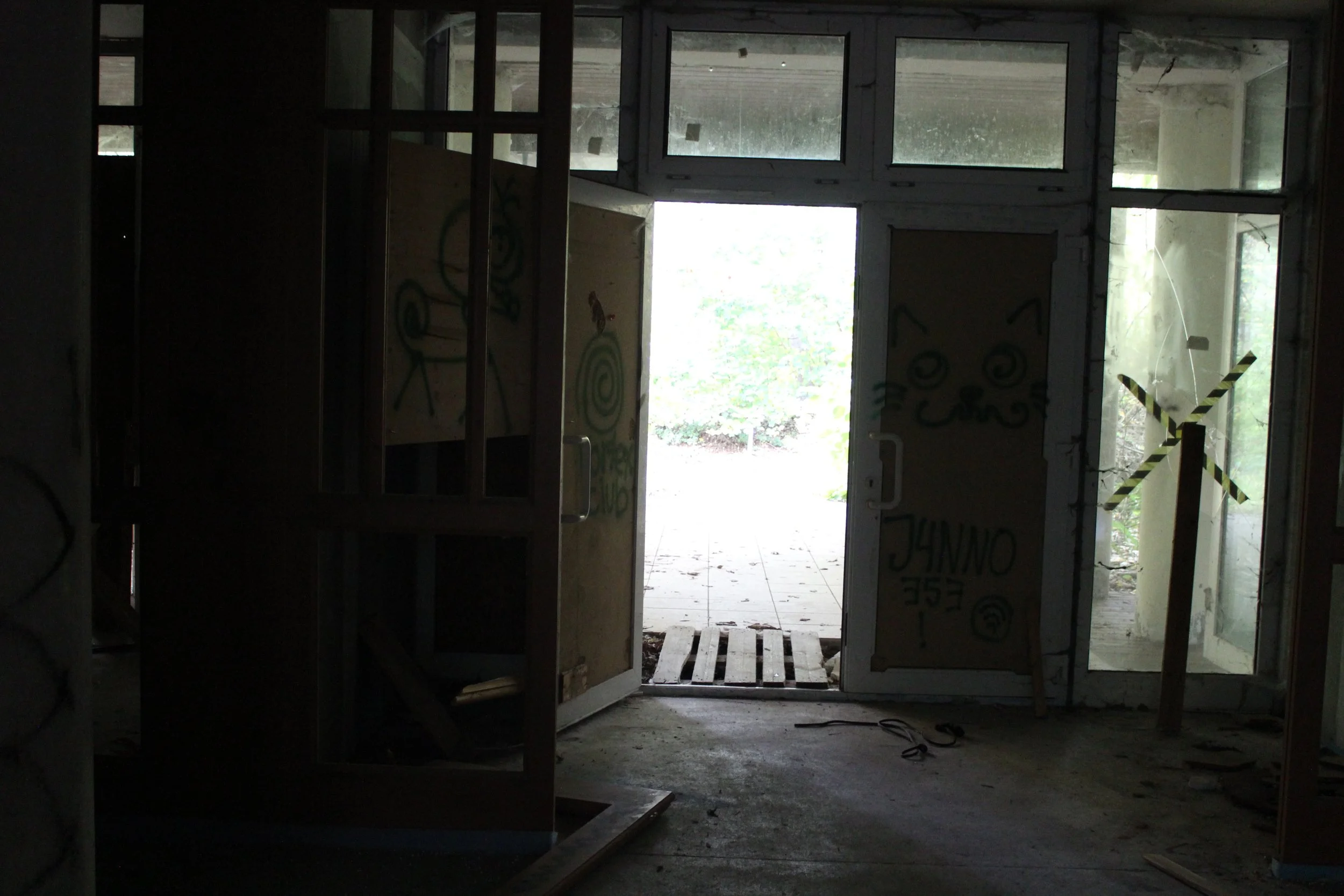 Innenansicht eines verlassenen, unvollendeten Gebäudes mit offenen Türen, durch die man ins Freie schaut. Die Türen sind mit Graffiti beschmiert. Das Licht kommt von draußen, während der Innenraum dunkel ist.
