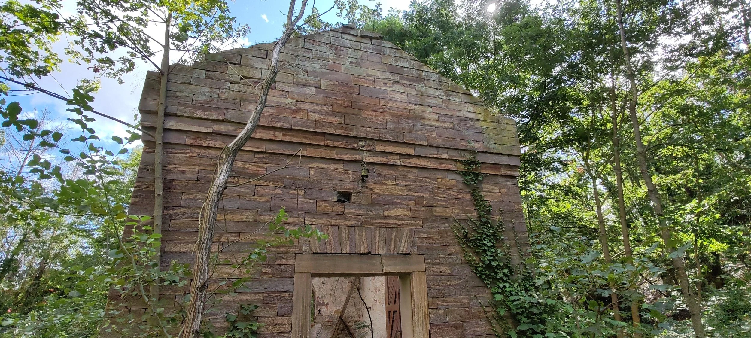 Verfallene, steinerne Ruine eines Gebäudes in einem dichten grünen Wald, am Tag, mit klarer blauer Himmel