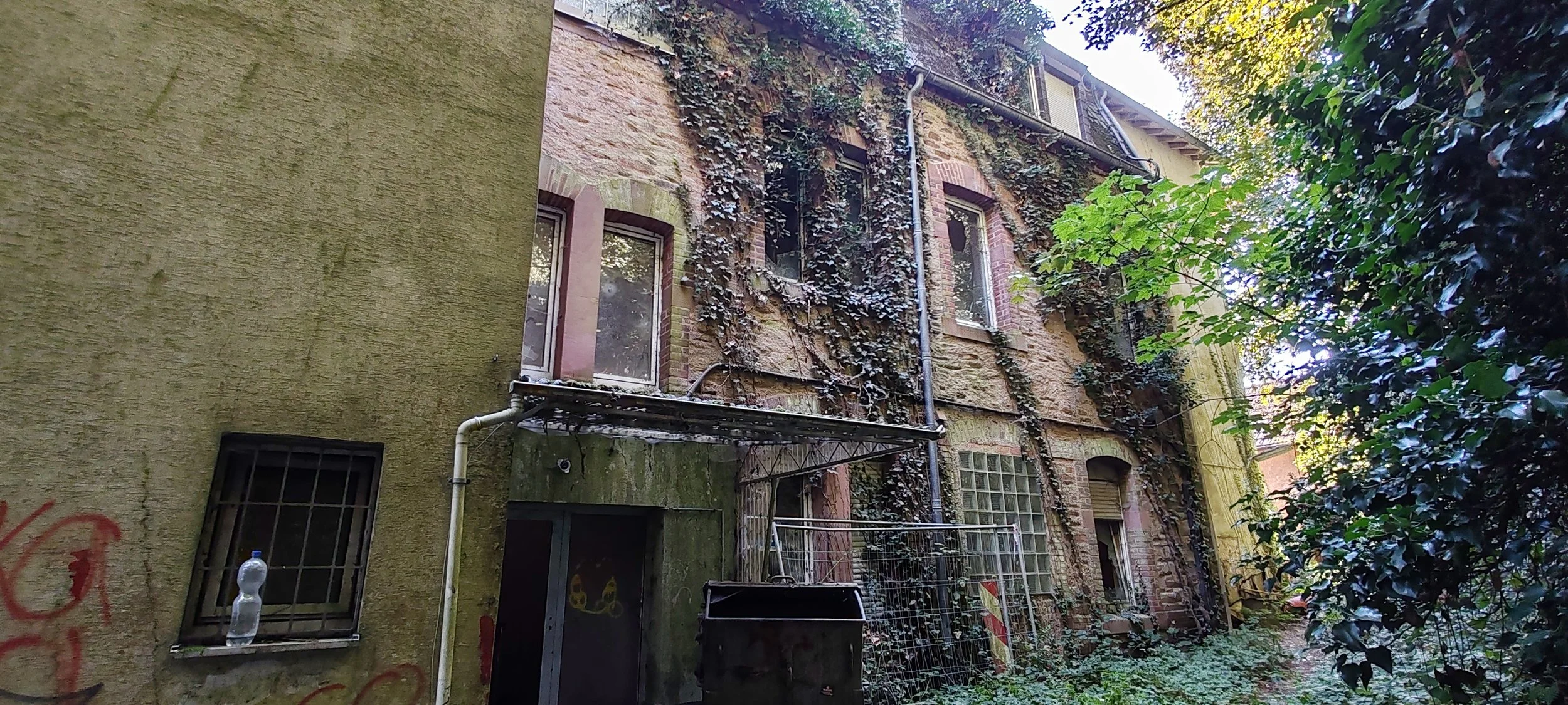 Ein altes, verfallenes Gebäude mit bröckelnder Fassade, überwuchert von Efeu und umliegendem Unkraut. Es gibt mehrere Fenster, einige mit Gittern, und einen Eingang. Das Bild wurde in einem bewachsenen, von Bäumen umgebenen Bereich aufgenommen.