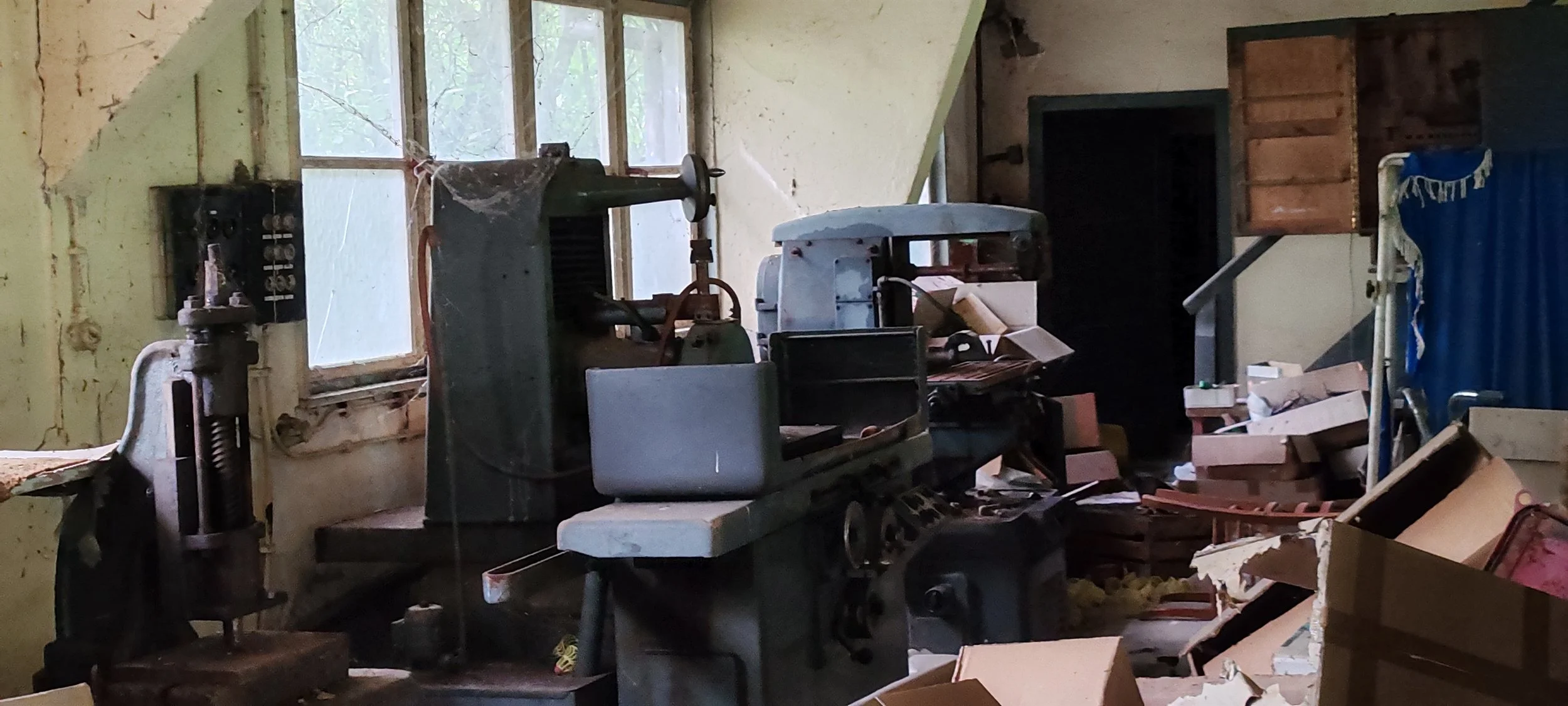 Ein verlassener Raum voller alter, verstaubter Werkzeuge und Maschinen, während die Fenster durch Staub und Spinnweben verziert sind, und Kartons und Holzreste umherliegen.