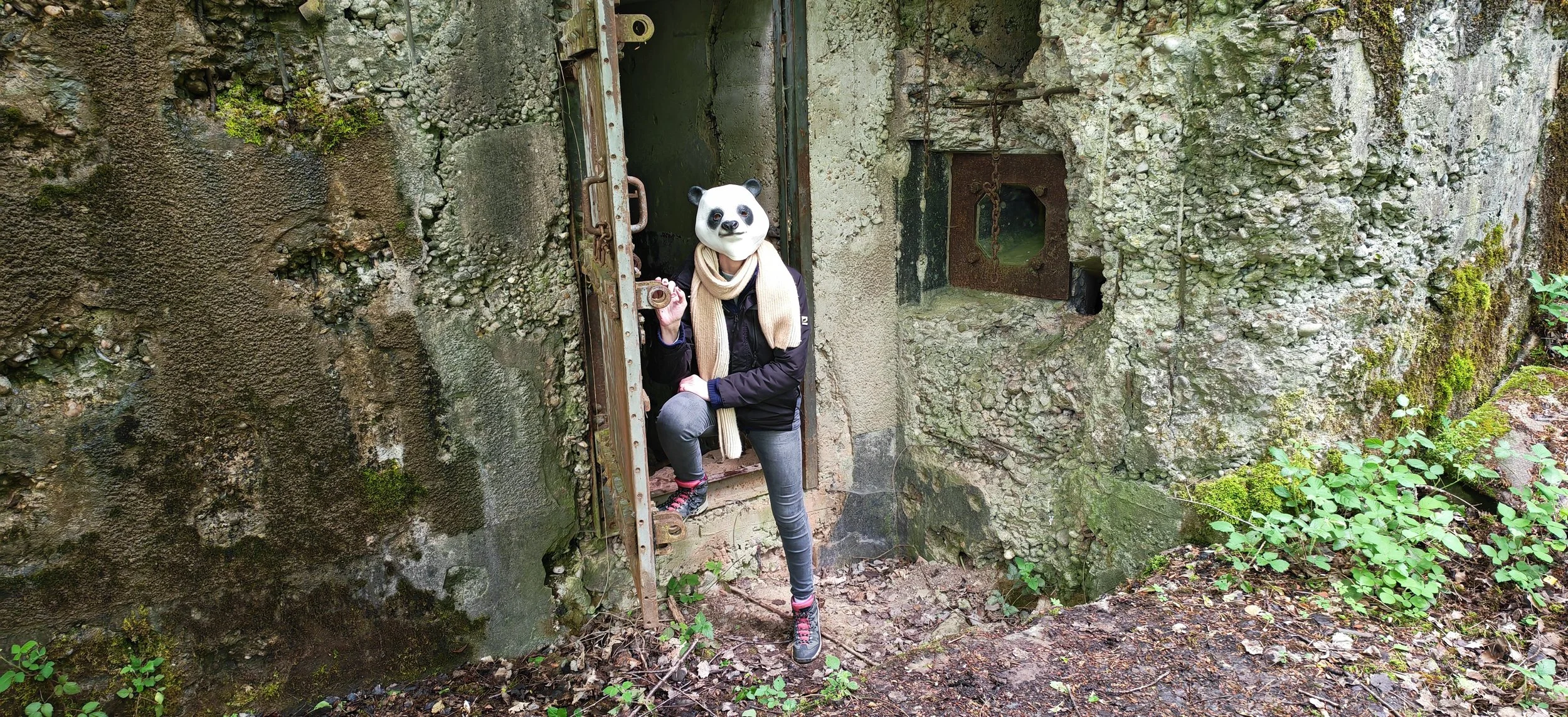 Person mit Panda-Maske steht an einer alten, moosbedeckten Tür aus Stein und Metall, umgeben von grüner Vegetation.