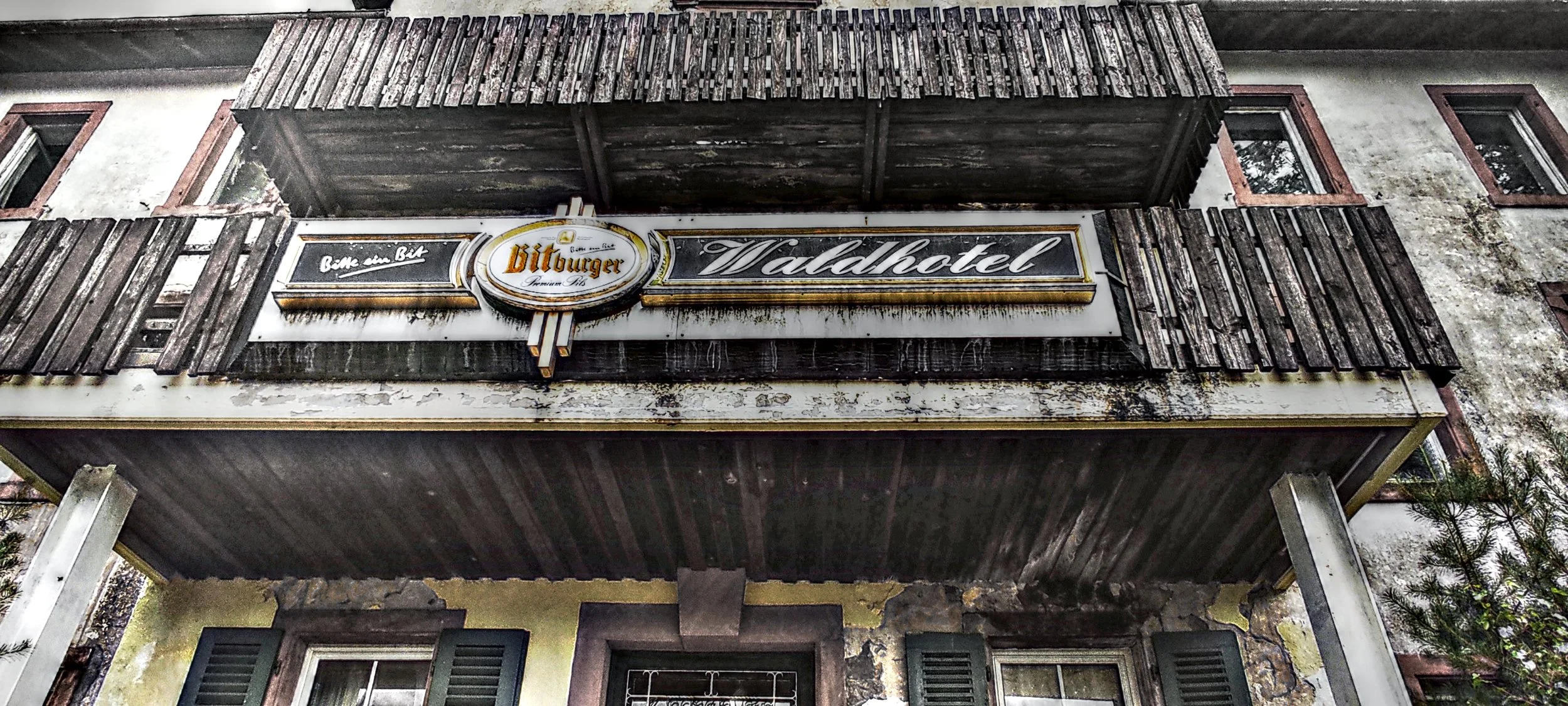 Blick auf das alte Hotel Valadhotel mit einem verblassten, verschmutzten Schild, das den Namen zeigt, und einem Balkon mit schiefen, schmutzigen Holzpanelen vor einem verputzten Gebäude mit zwei Fenstern.