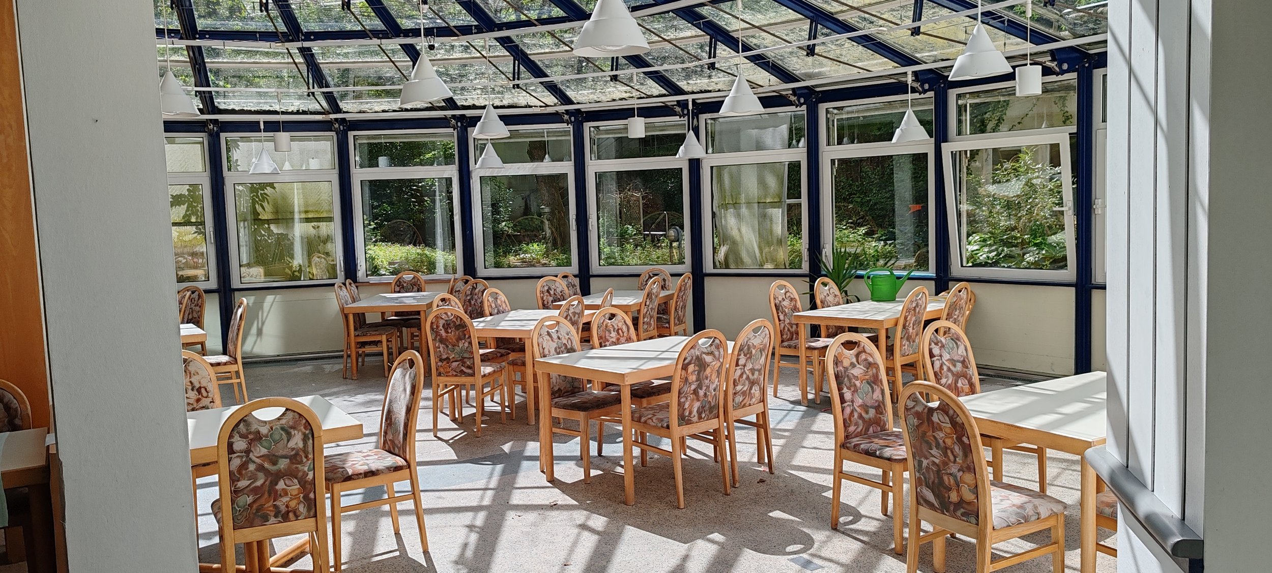 Ein heller Wintergarten mit mehreren Tischen und Stühlen, Sonnenlicht scheint durch die Dachfenster, Pflanzen auf den Tischen.