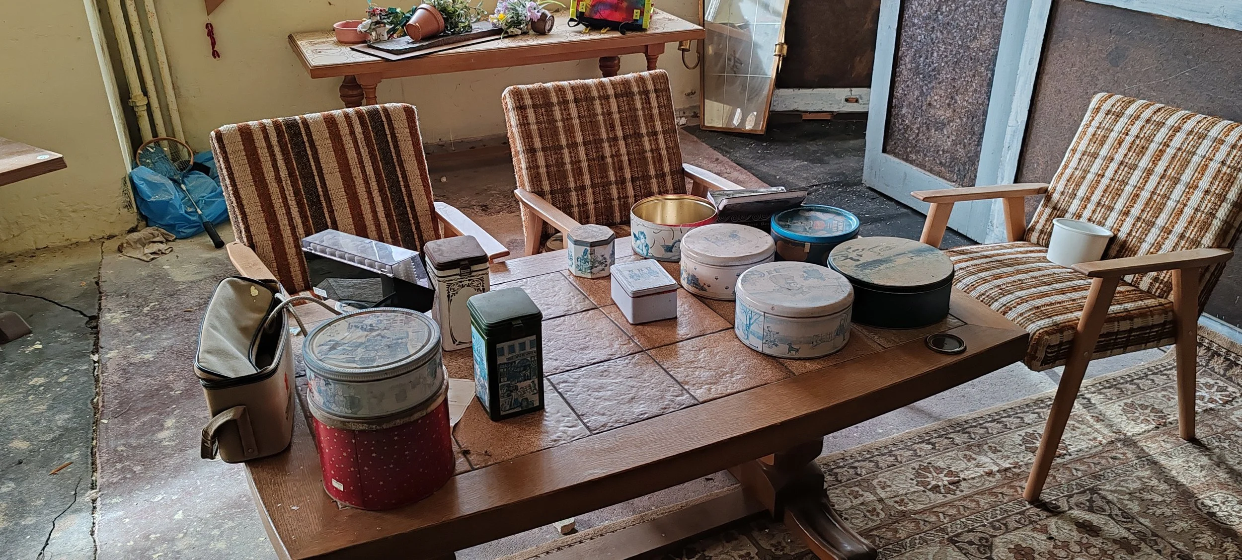 Ein Holztisch mit mehreren bunten Aufbewahrungsboxen, Tassen und anderen Gegenständen. Im Hintergrund ein Spiegel und eine Tür, umgeben von einer ungepflegten Umgebung mit Teppich und rohem Boden.